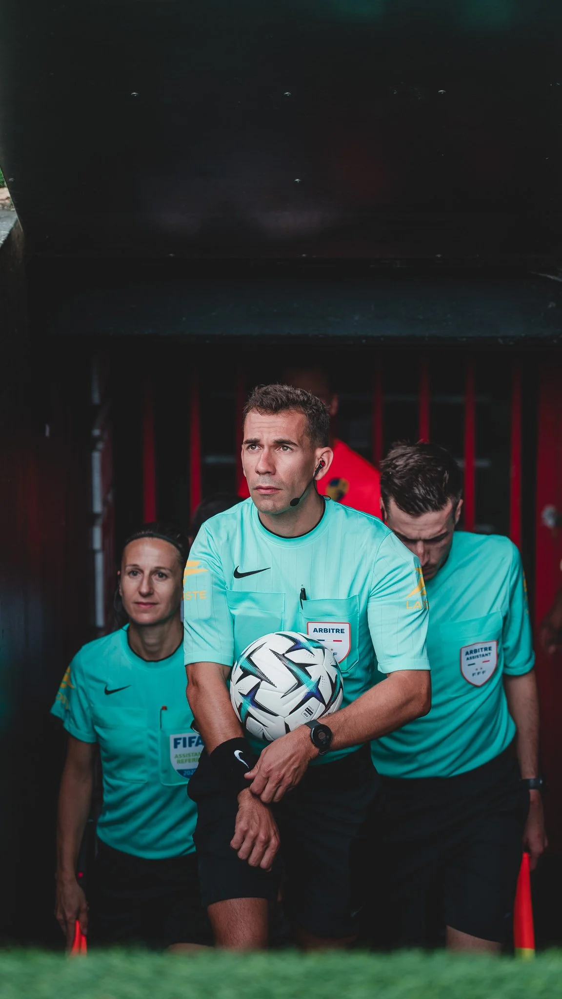 Arbitre de football tenant un ballon, accompagné de deux assistants, tous portant des maillots bleus et marchant dans un tunnel avant un match.