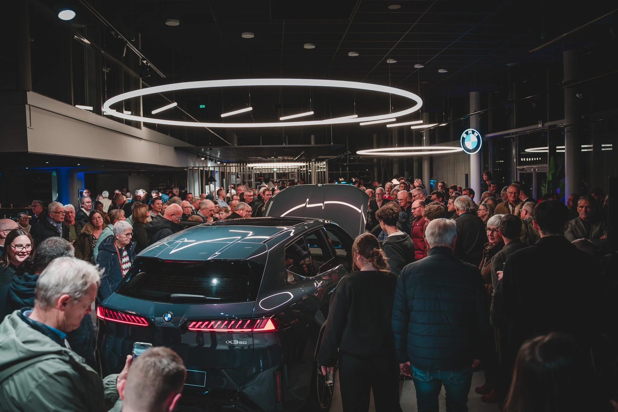 Une foule de personnes dans un salon auto, entourant une voiture BMW noire, avec un logo BMW visible sur le mur à droite.