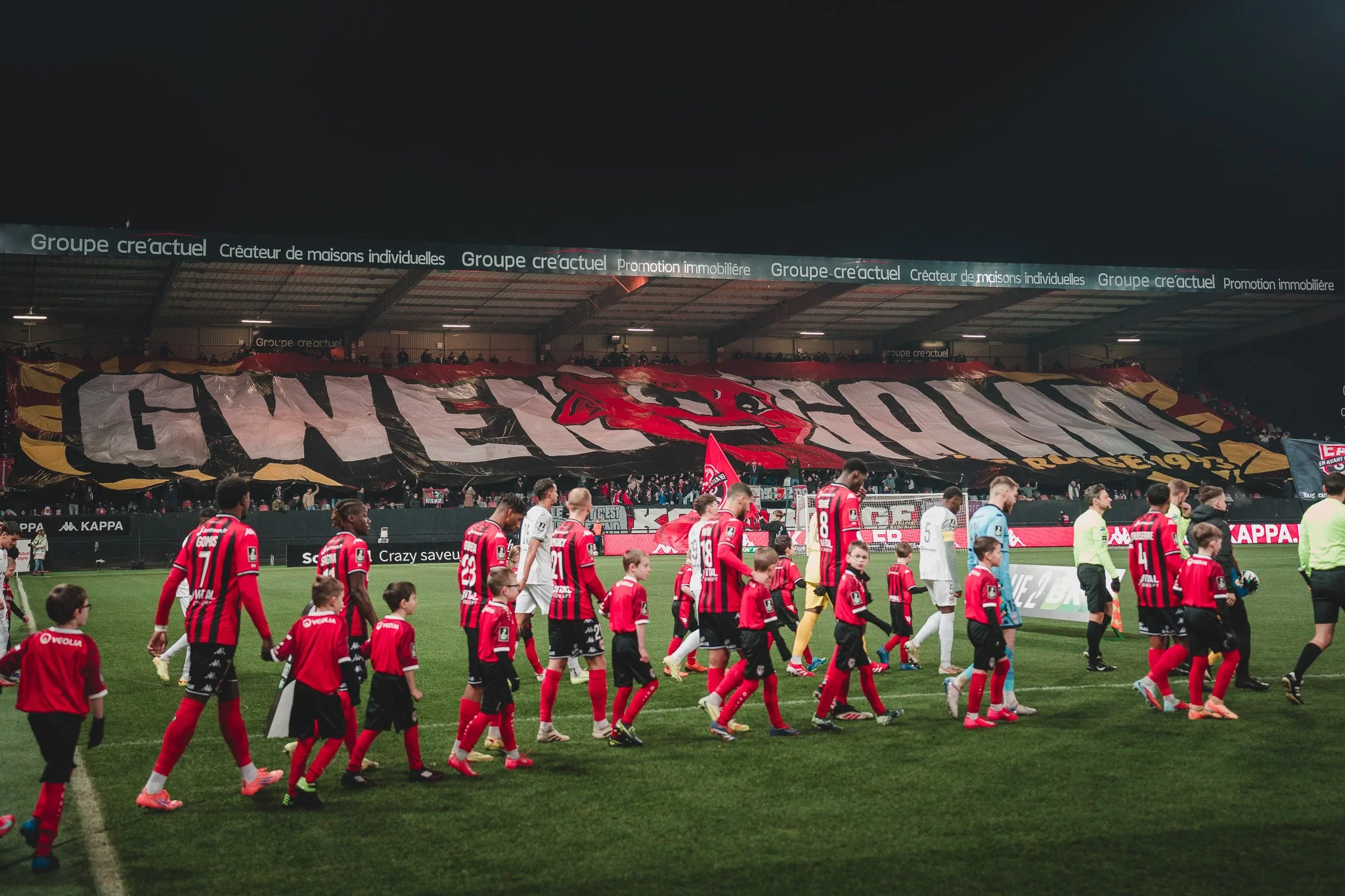 Des joueurs de football, accompagnés de jeunes enfants, marchent sur un stade en soirée. Un grand drapeau avec le logo du club, probablement le Stade Malherbe de Caen, est déployé en arrière-plan dans les gradins. La scène est lumineuse avec des publicités sur le pourtour du terrain.
