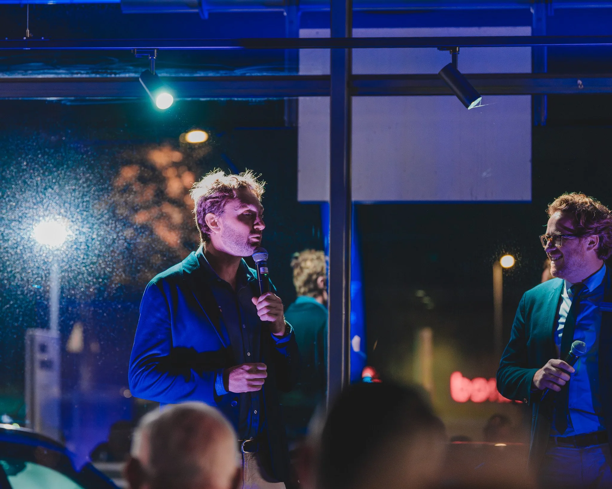 Deux hommes en costume discutent avec des microphones dans un lieu sombre avec des lumières colorées, probablement lors d'une présentation ou d'un événement en soirée.