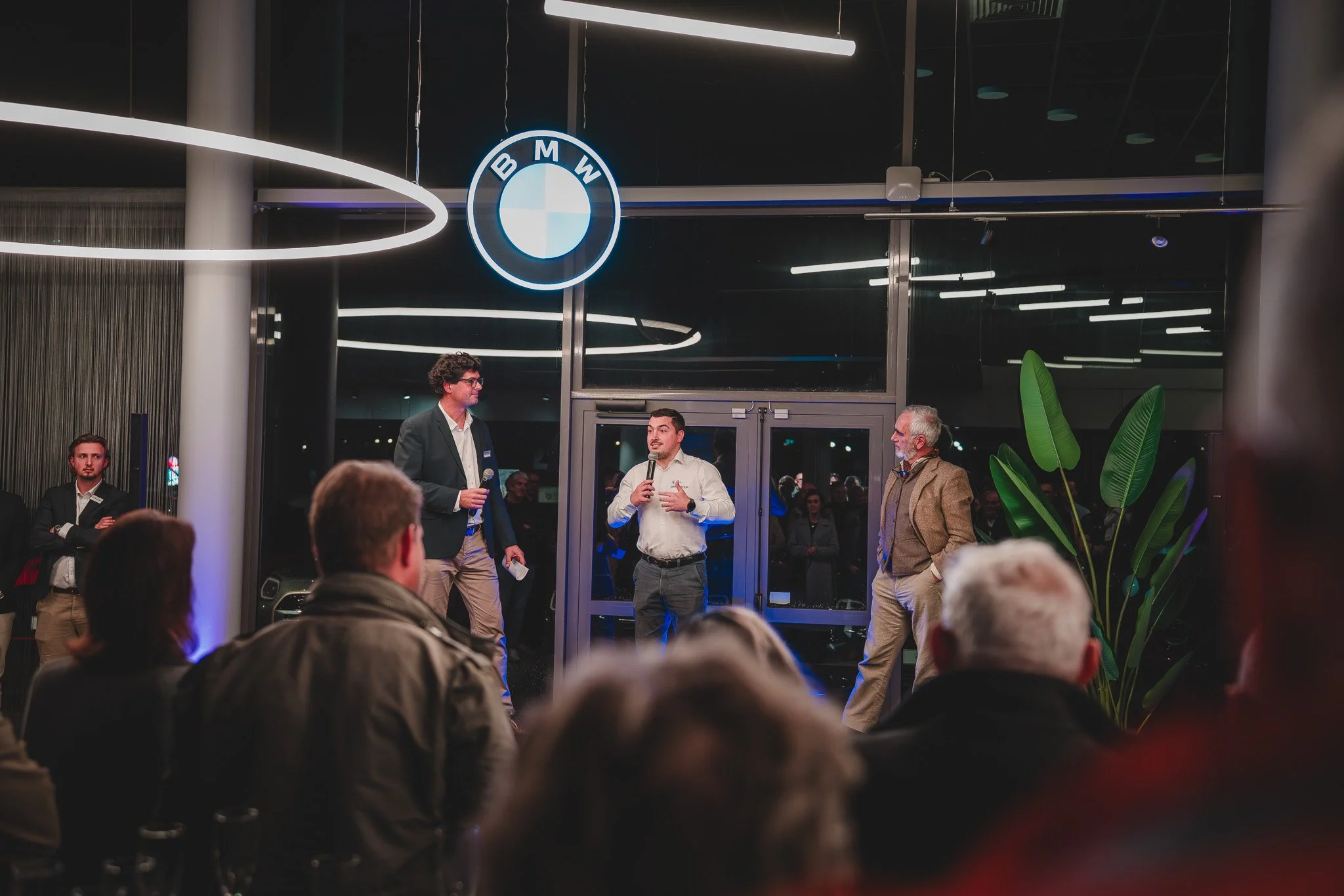 Une conférence ou un événement professionnel avec des intervenants parlant à un public, dans un endroit moderne avec un logo BMW illuminé au plafond.