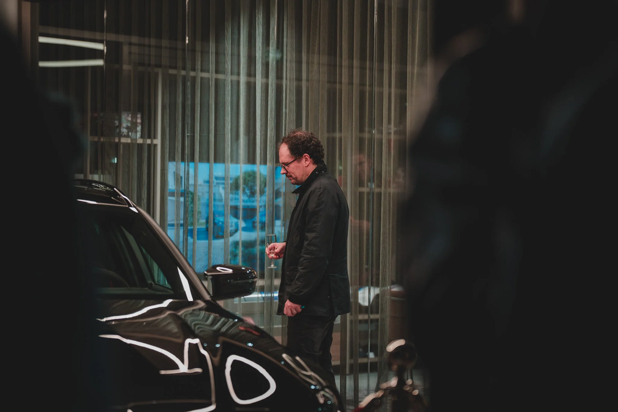 Un homme portant un costume noir et des lunettes, tiendra un verre, se tient près d'une voiture noire dans une salle d'exposition ou un garage, avec des rideaux en arrière-plan.