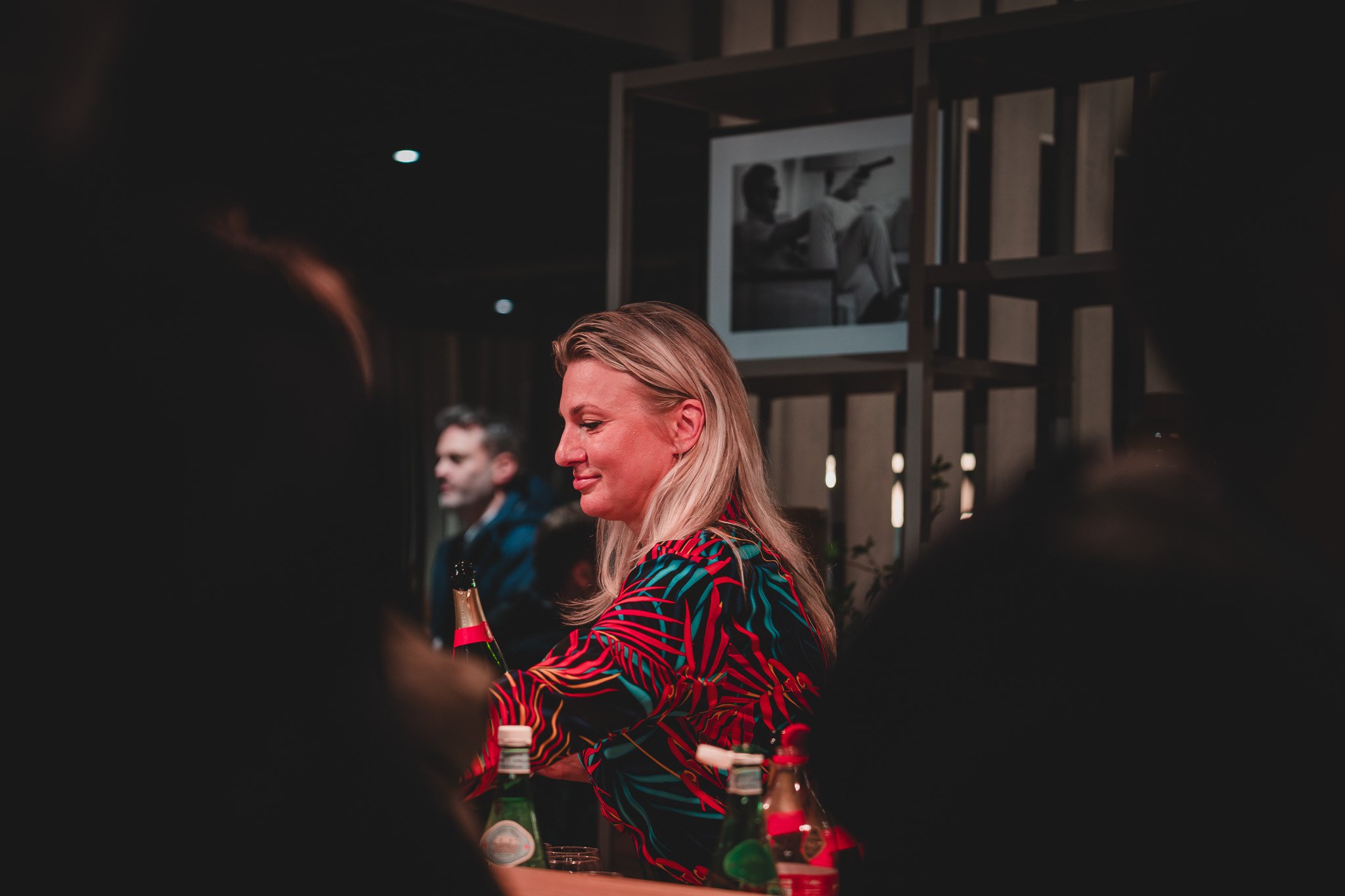 Une femme blonde souriante assise à un bar ou une table, entourée de bouteilles de boisson, dans un environnement intérieur chaleureux et feutré.