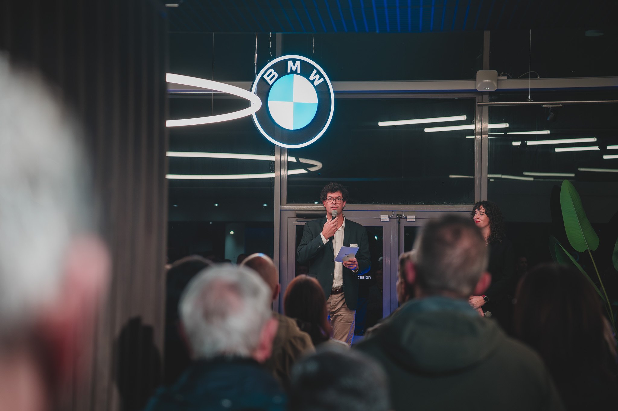 Un homme en costume parle lors d'une conférence dans une salle avec un logo BMW en arrière-plan.