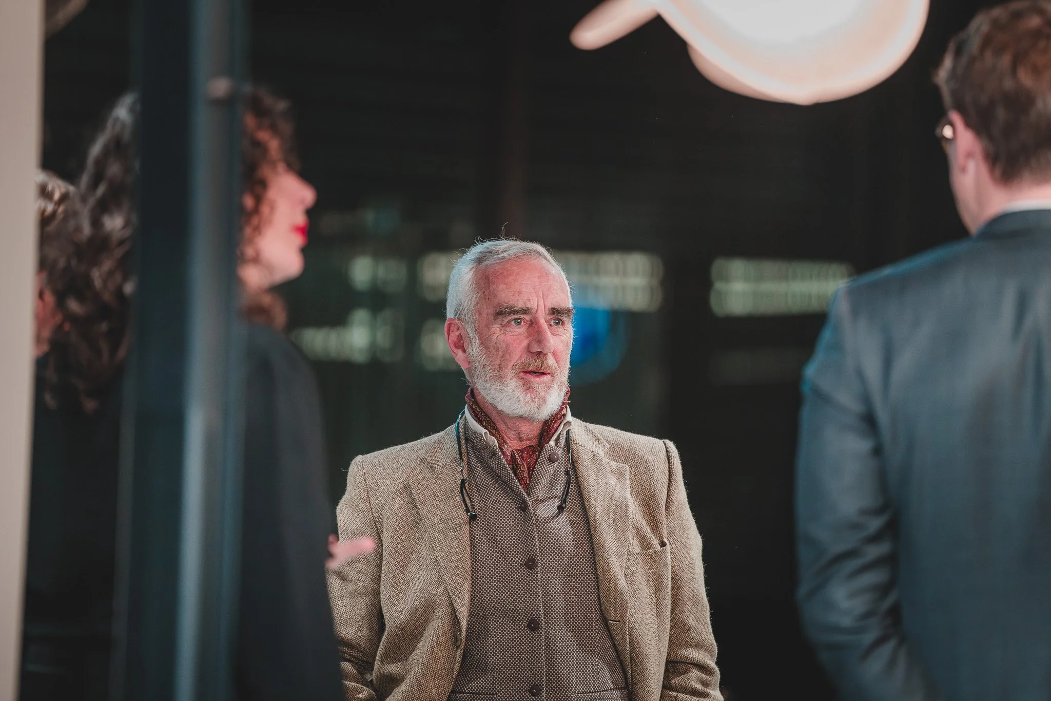 Trois personnes en conversation dans une pièce sombre, avec un homme âgé au centre portant un manteau beige, une femme à gauche avec des cheveux bouclés, et un jeune homme à droite avec des lunettes et un costume