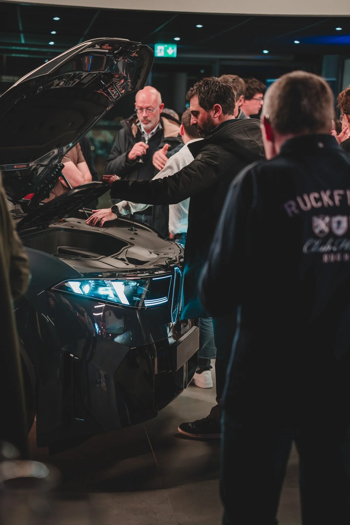 Groupe de personnes autour d'une voiture noire avec le capot levé, dans un environnement intérieur, probablement une exposition ou un salon automobile.