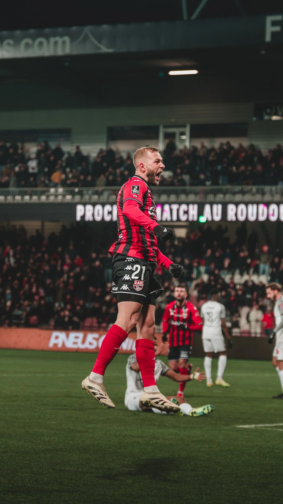 Un joueur de football en maillot rouge et noir célèbre sur le terrain lors d'un match, avec des spectateurs dans les tribunes en arrière-plan.