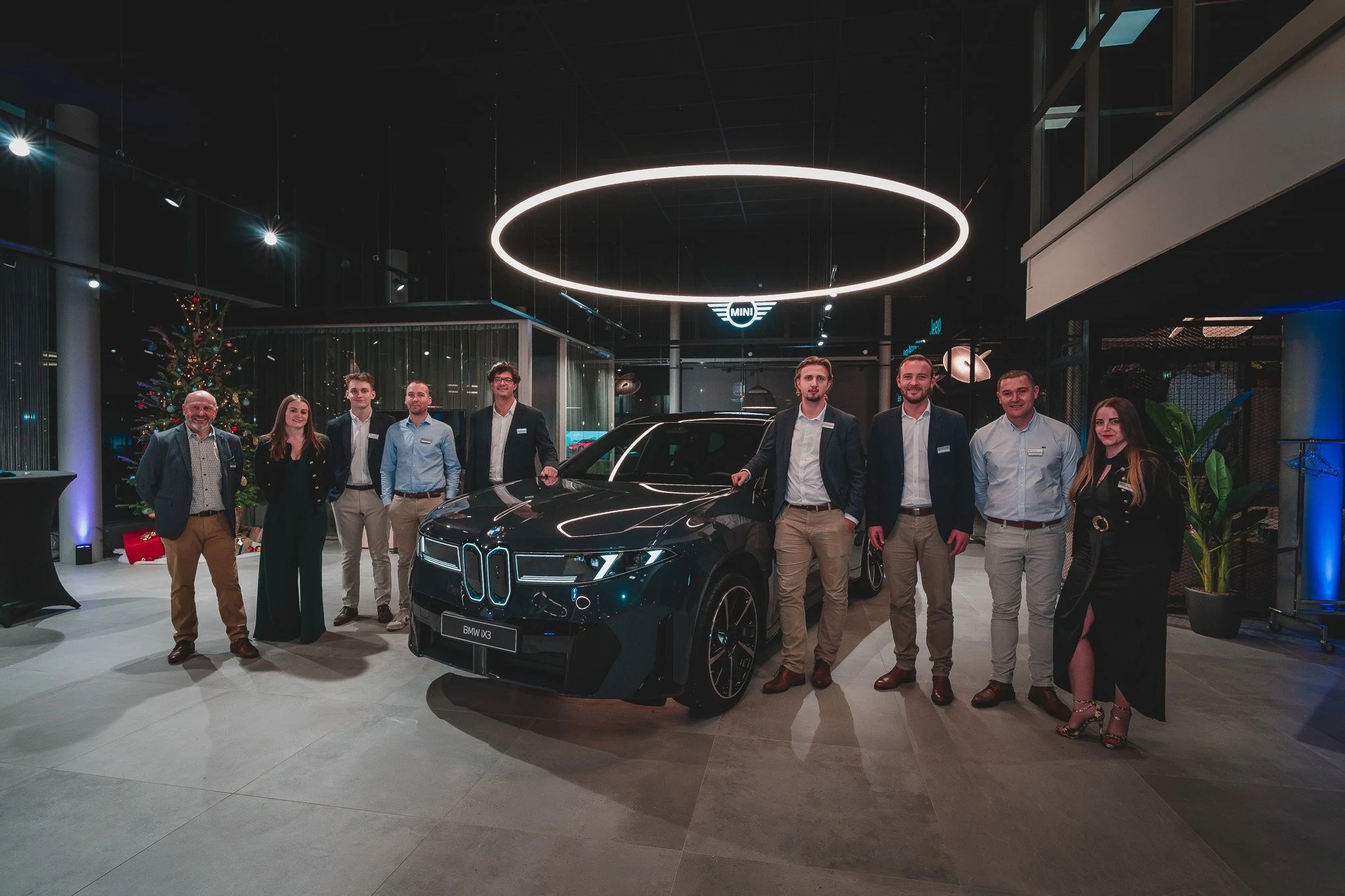 Groupe de personnes debout autour d'une voiture noire électrique dans un showroom, décoré pour Noël, avec un arbre de Noël en arrière-plan, sous un plafonnier circulaire lumineux.