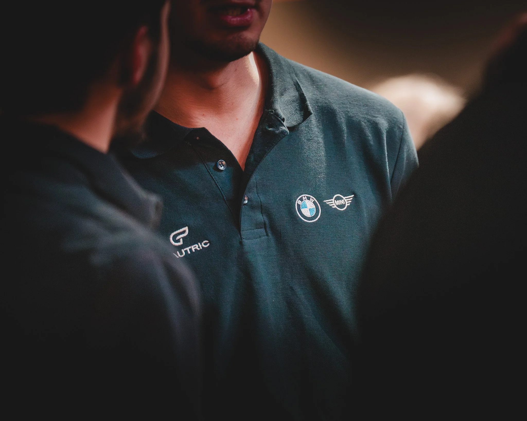 Gros plan d'une personne portant un polo avec les logos BMW, MINI, et UTTRIC, discutant avec d'autres personnes.