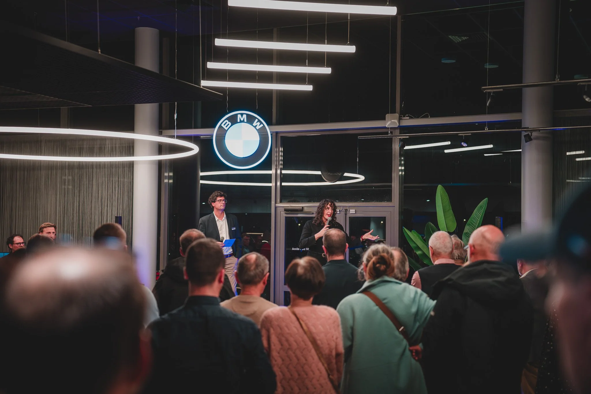 Une réunion ou une présentation dans un espace moderne avec un grand groupe de personnes. Deux personnes sont sur scène, l'une parlant avec un micro et l'autre tenant un document, avec le logo de BMW derrière eux. La salle est éclairée par des éclair