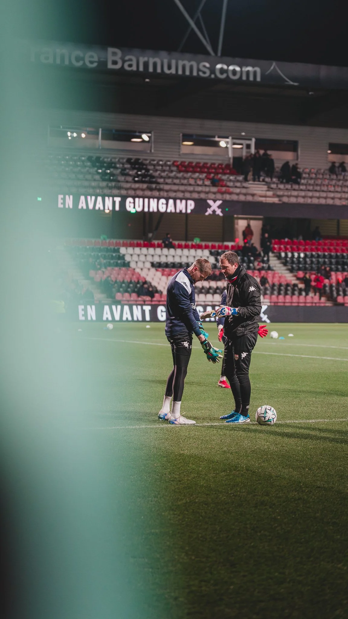 Deux gardiens de but discutent sur un terrain de football avant un match, entourés de ballons et dans un stade vide.