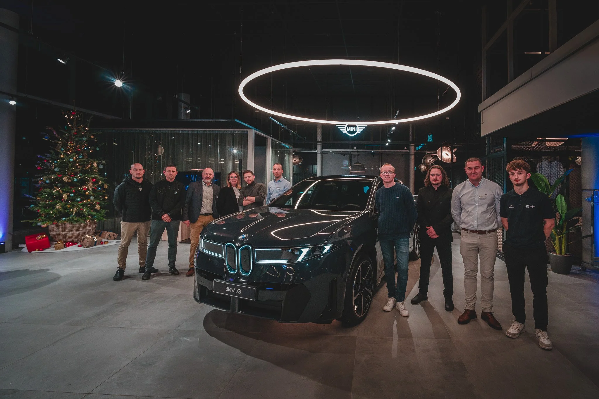 Groupe de personnes posant autour d'une voiture BMW iX3 dans un showroom, décoré avec un sapin de Noël, à l'intérieur d'un bâtiment moderne.