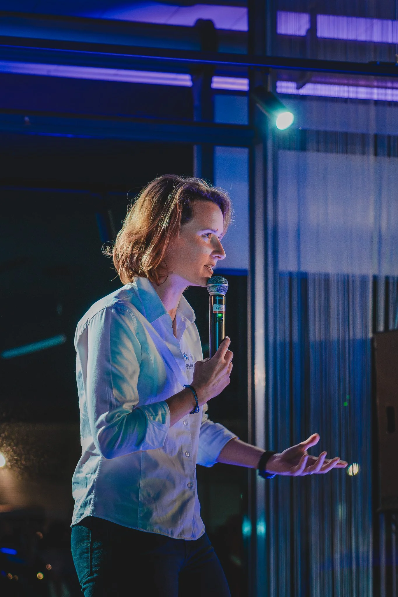 Une femme parle devant un microphone lors d'une présentation ou conférence, sous un éclairage violet et bleu.