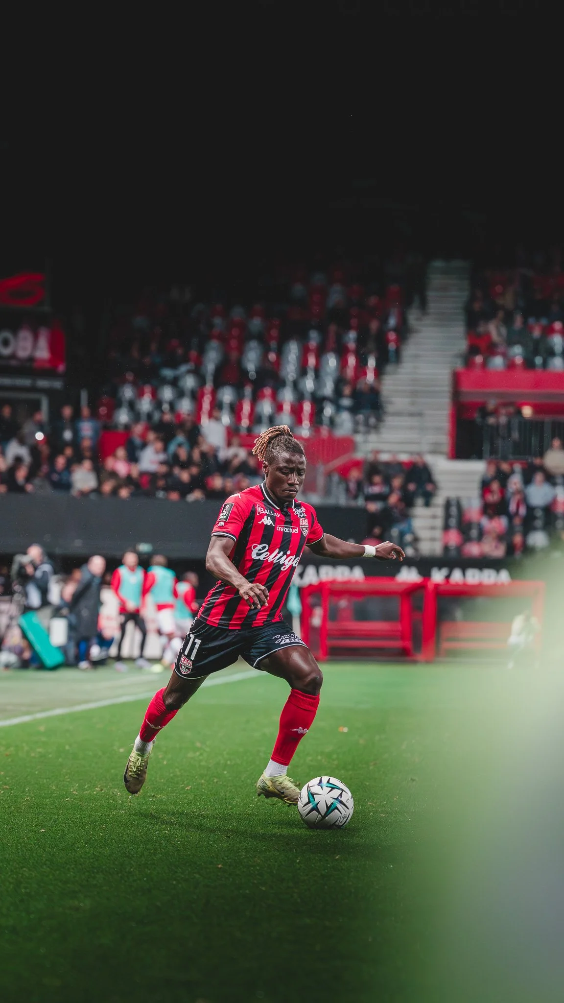 Un joueur de football en maillot rouge et noir en action sur le terrain, avec le public en fond.
