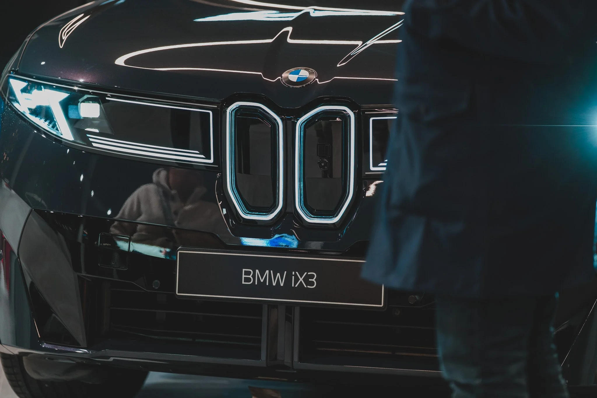 Première voiture BMW iX3 avec un logo BMW visible, montrant le devant noir et les phares modernes, à l'intérieur d'une salle d'exposition.