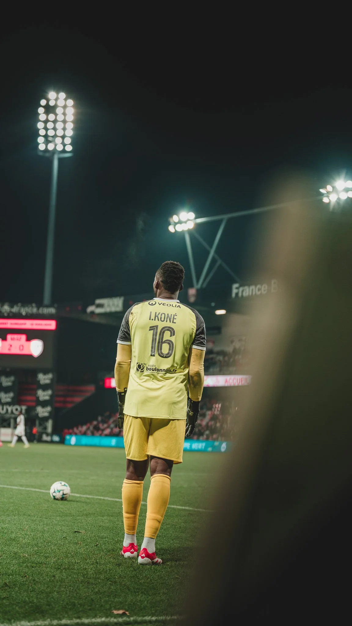 Un joueur de football masqué, vêtu d'un short et d'un maillot jaune portant le nom I. KONÉ et le numéro 16, se tient sur le terrain lors d'un match nocturne.