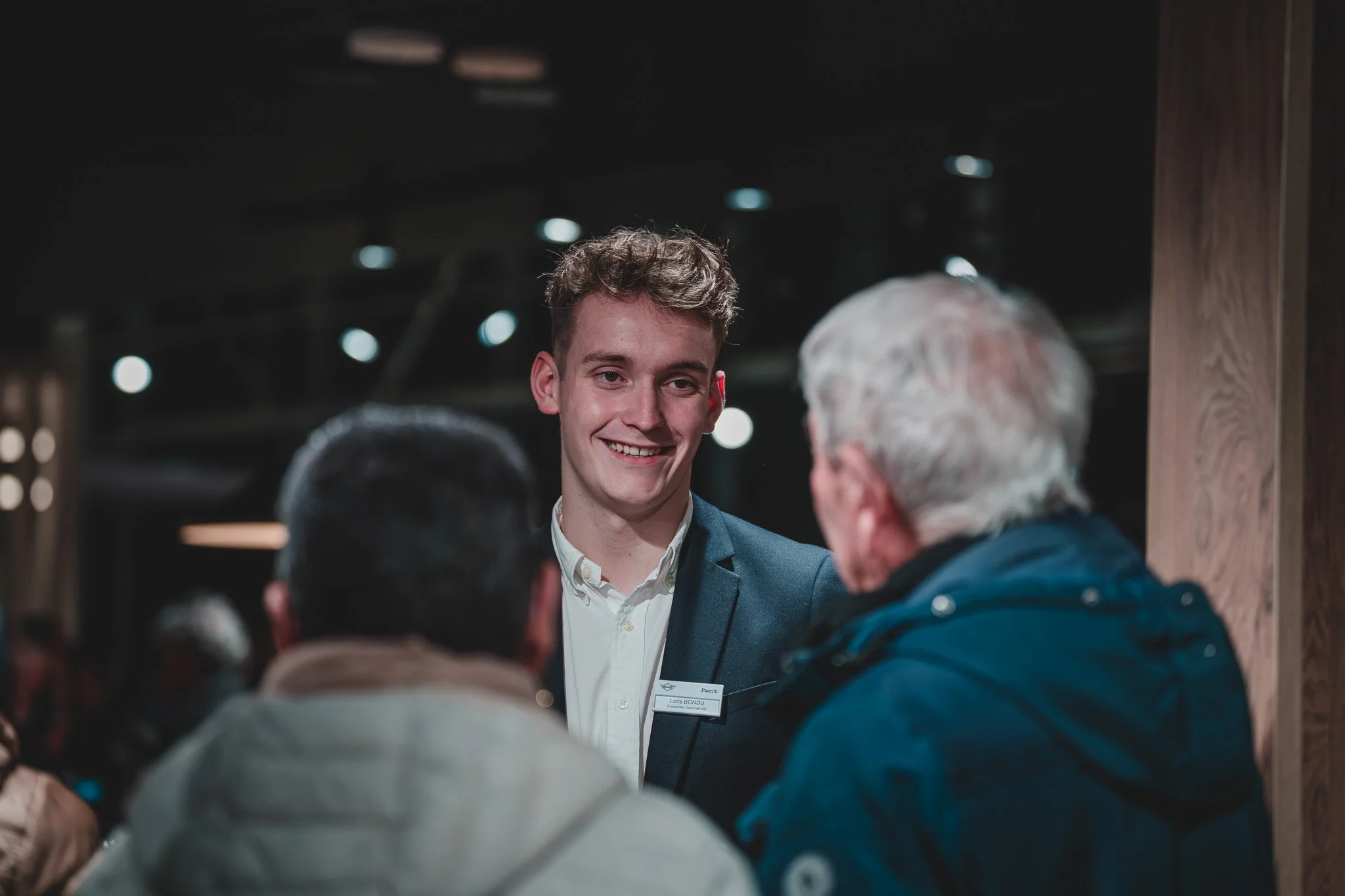 Un jeune homme souriant en costume parle à deux personnes plus âgées dans un lieu intérieur, probablement lors d'un événement ou d'une conférence.