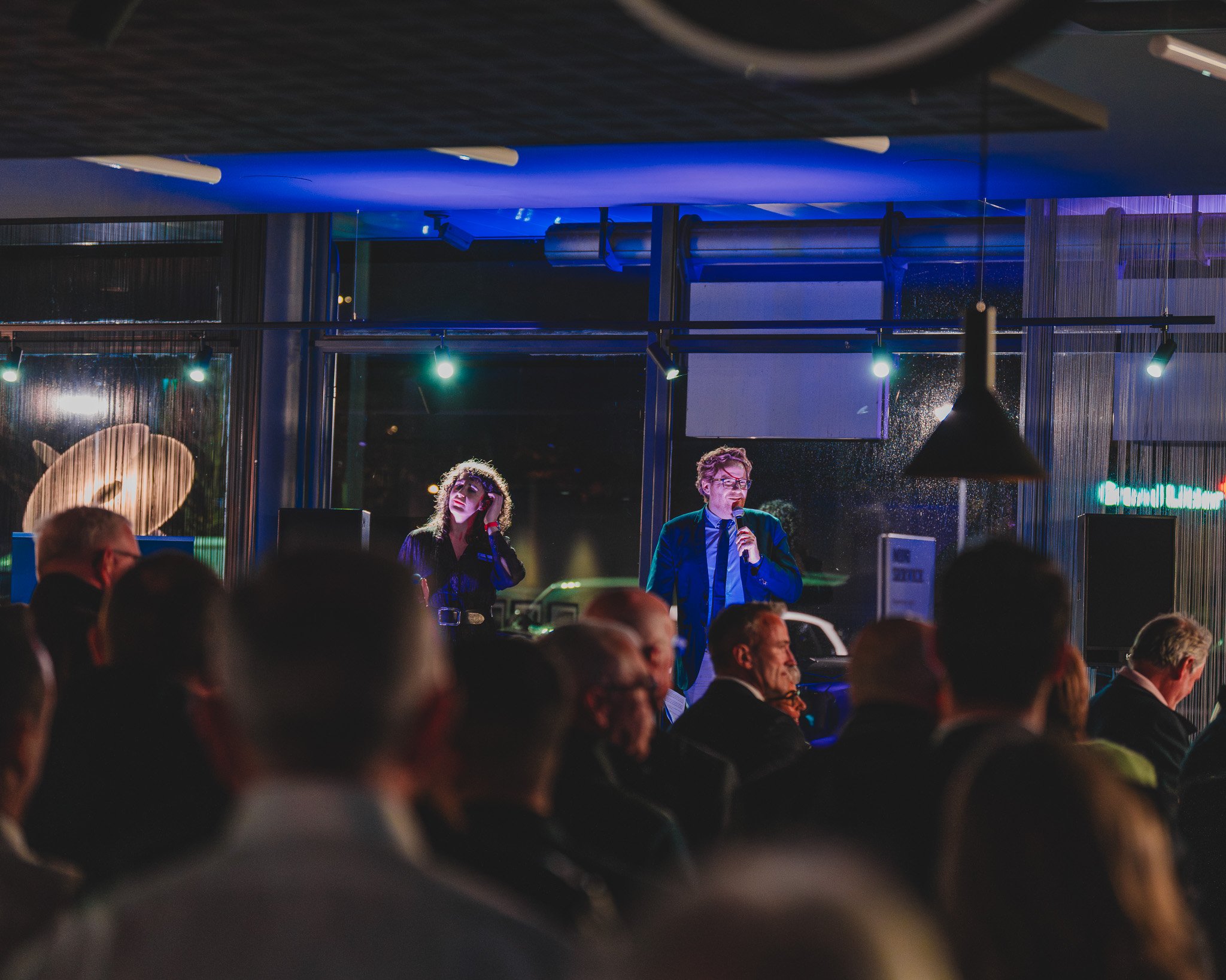 Une scène d'un événement ou d'une conférence avec un homme en costume parlant dans un microphone et une femme à côté avec un casque, tous deux sur une scène devant une audience assise dans une salle moderne avec grands fenêtres.