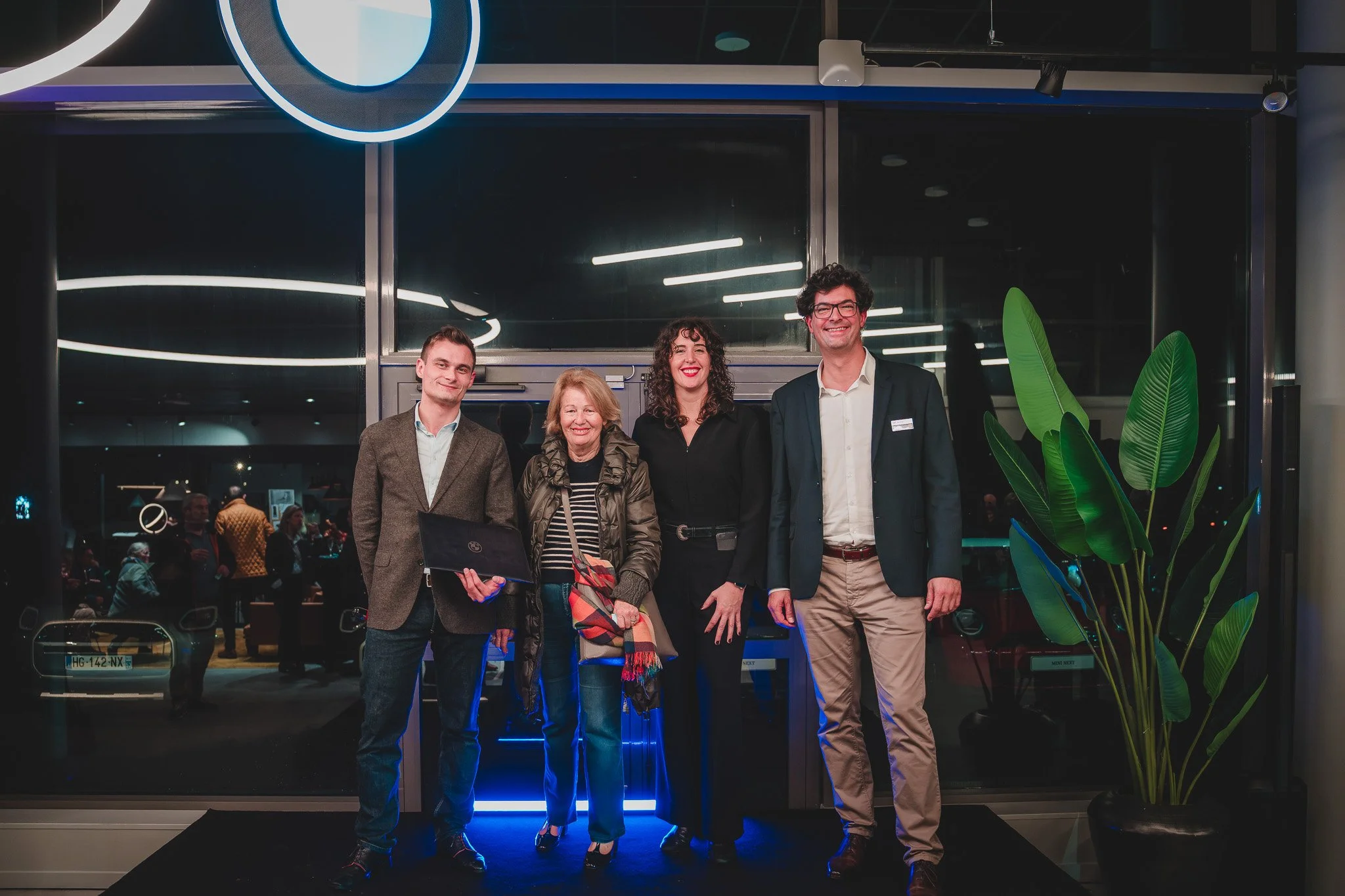 Groupe de quatre personnes posant devant une entrée, avec un grand plante verte à droite. C'est une scène en intérieur à l'éclairage moderne.