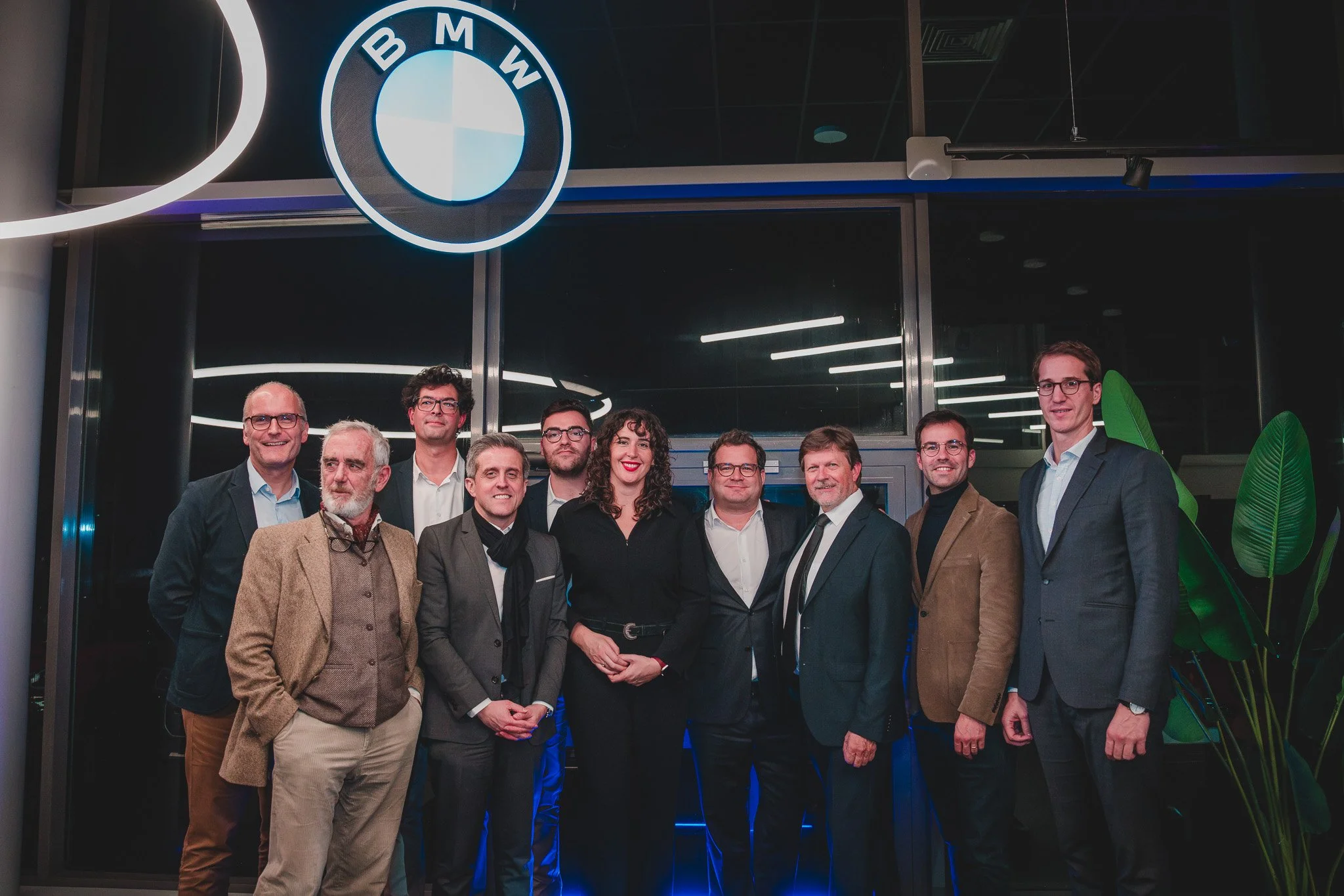 Groupe de personnes posant devant un mur en verre avec un logo BMW éclairé. Il y a des hommes et une femme en costume professionnel, quelques plantes vertes à droite.