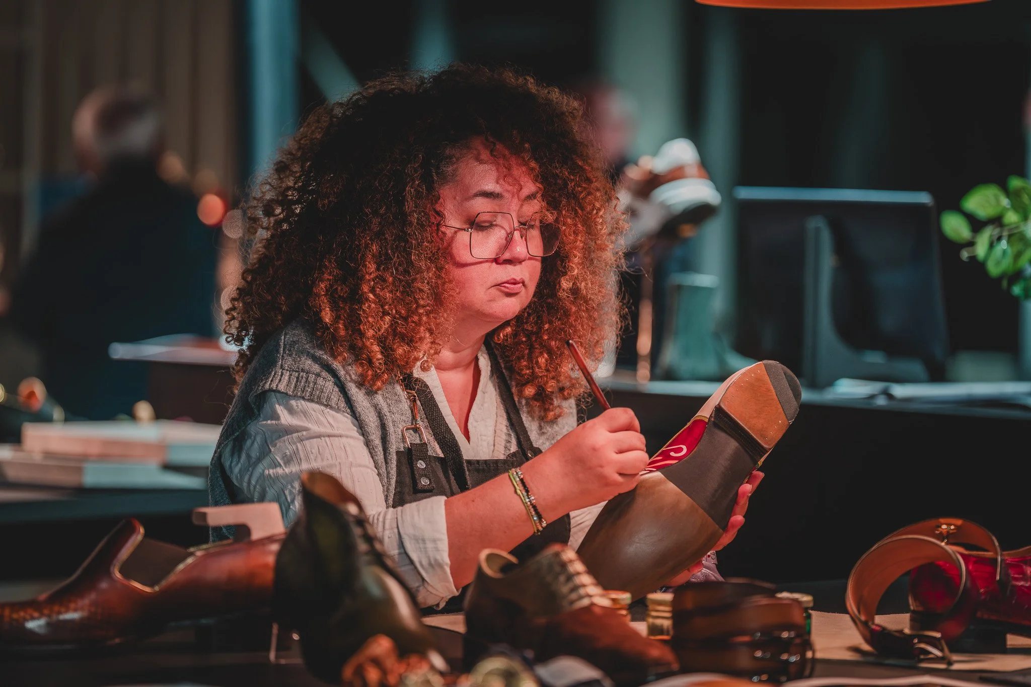 Femme avec cheveux frisés et lunettes, en train de peindre une botte en cuir dans un atelier ou magasin de chaussures.