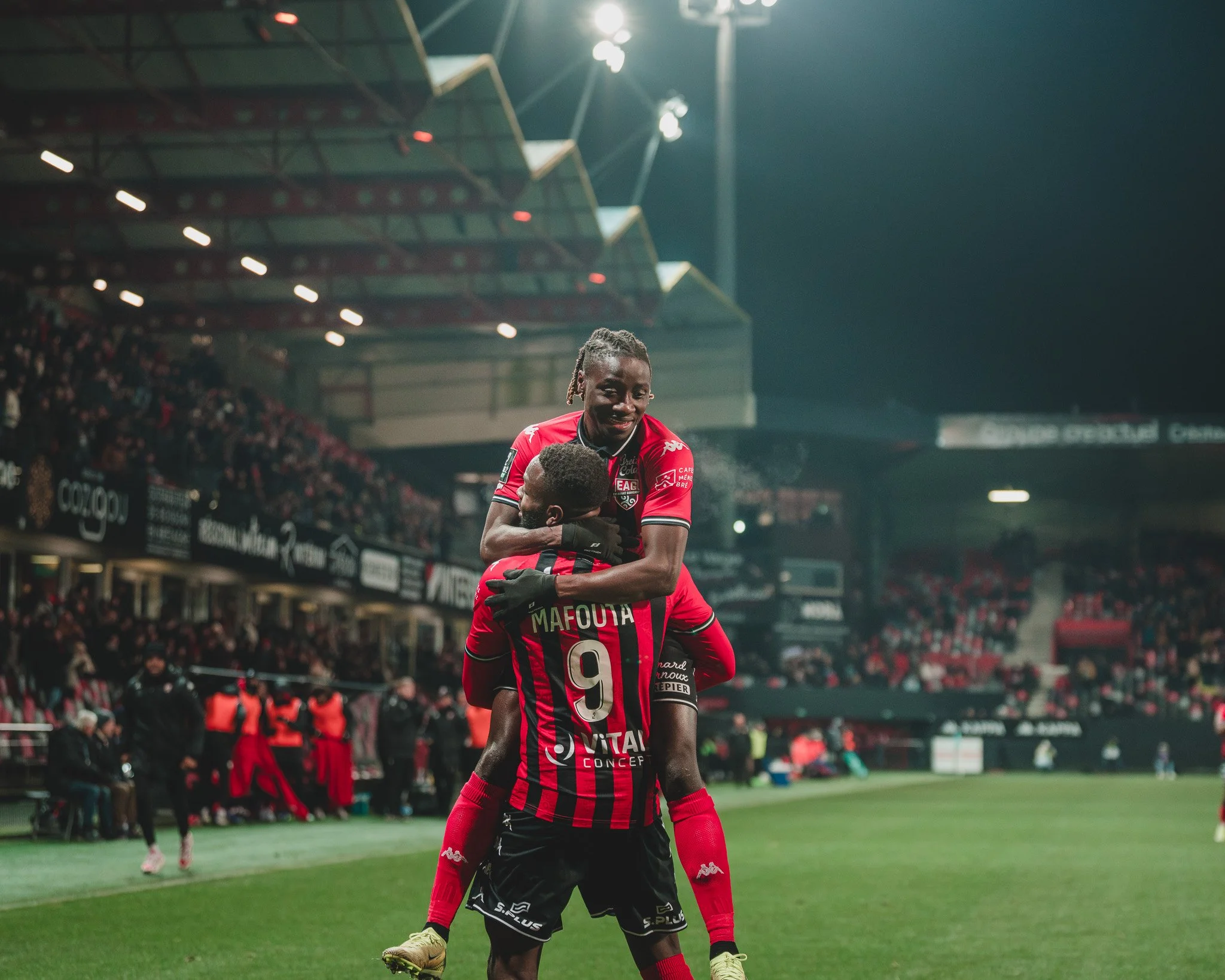 Deux joueurs de football en tenue rouge célébrant un but sur le terrain d'un stade, un saut en l'air et une étreinte