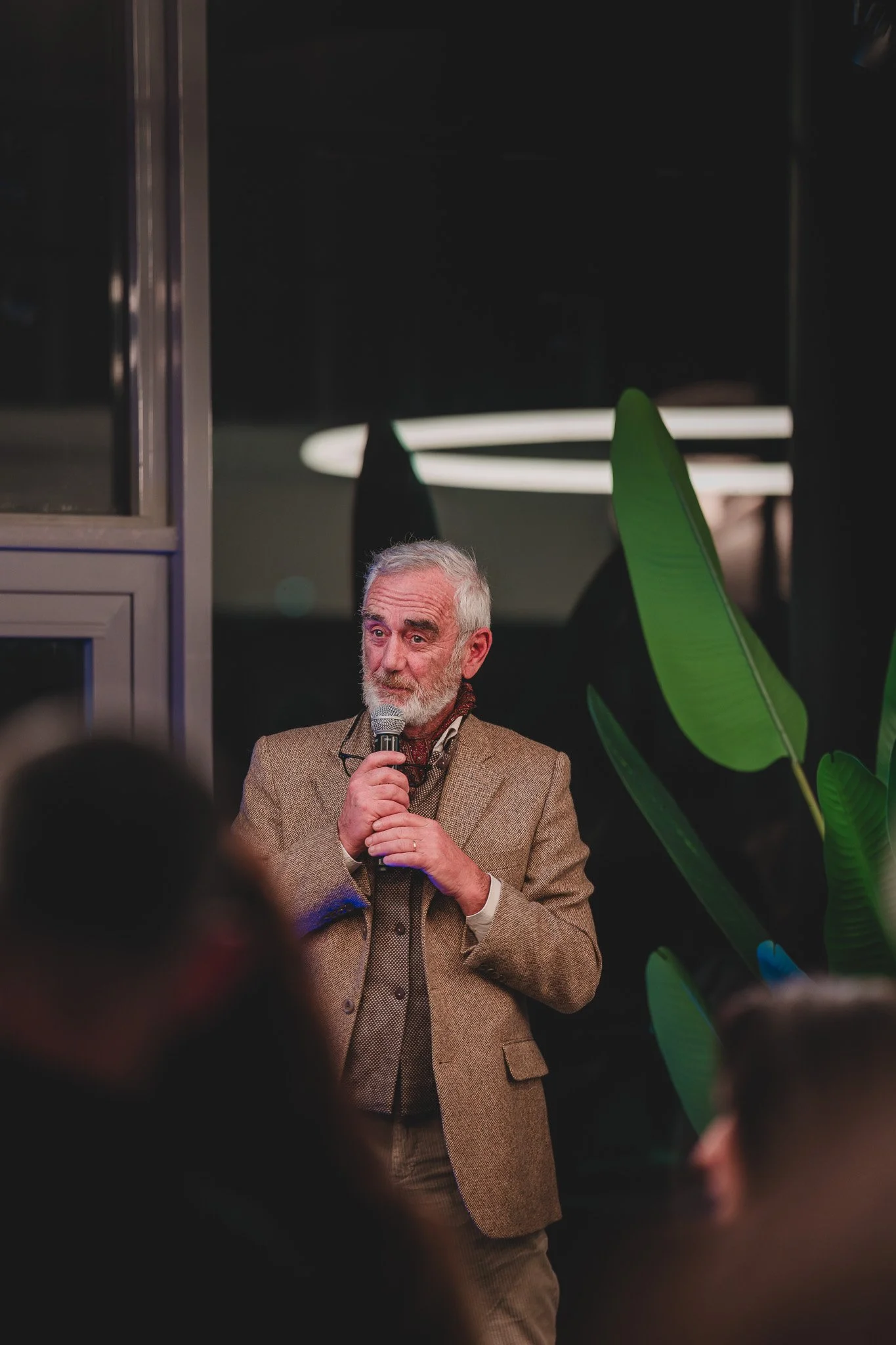 Un homme âgé, avec une barbe grise, portant un costume marron, parle dans un micro devant un groupe de personnes dans un cadre intérieur, avec des plantes vertes en arrière-plan.