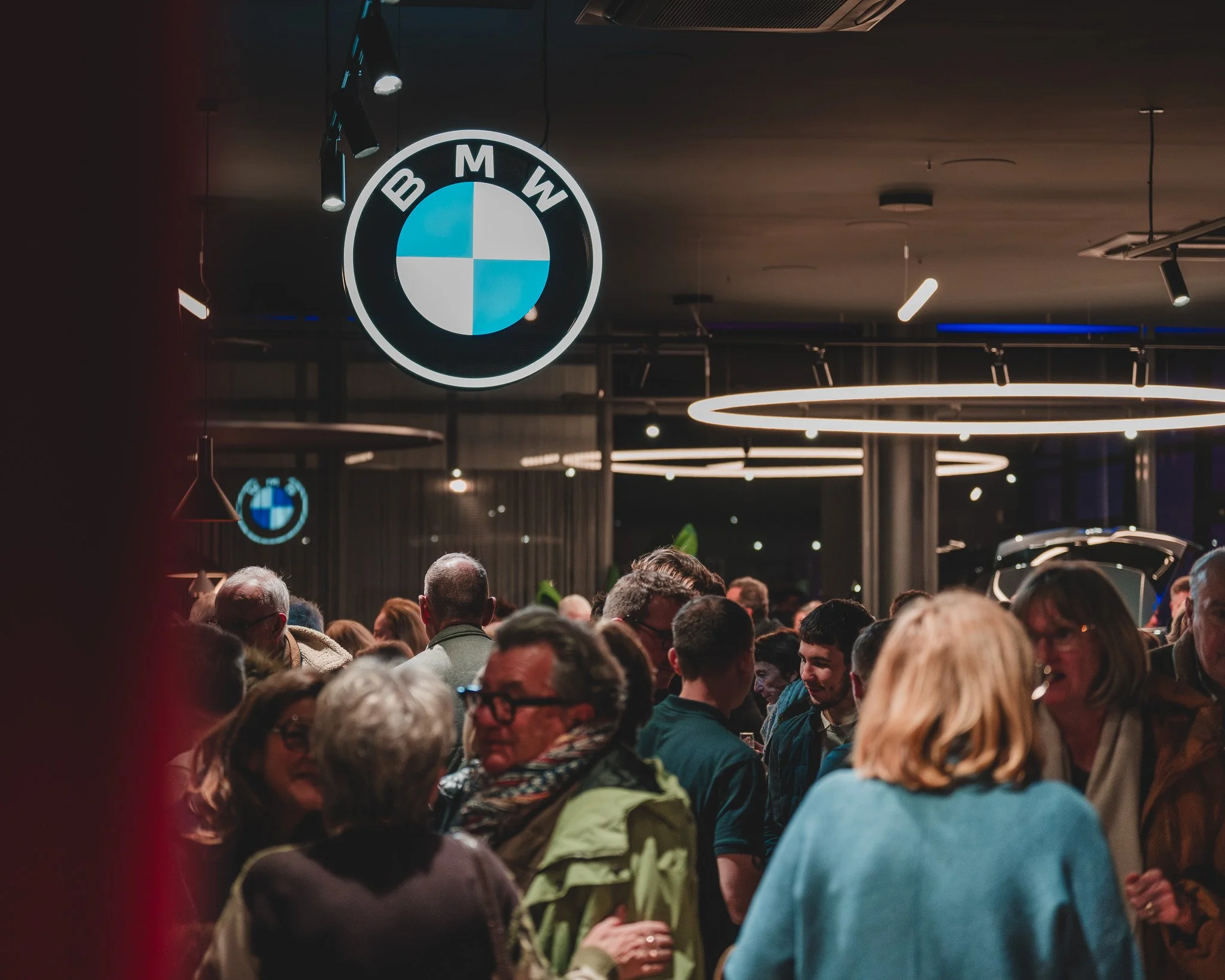Une foule de personnes dans un espace intérieur avec des logos BMW suspendus au plafond, ambiance lumineuse moderne.