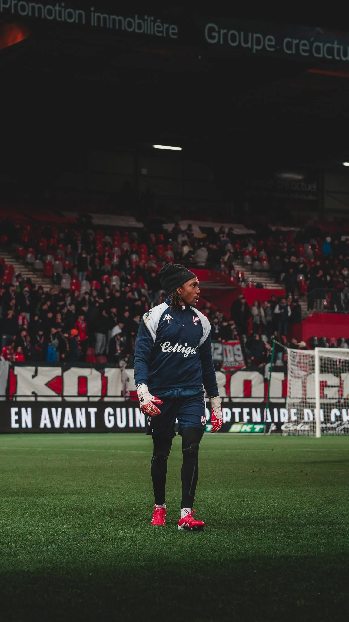 Un joueur de football sur un terrain, portant un costume d'entraînement avec des gants, sous un stade rempli de spectateurs, en arrière-plan un panneau publicitaire et des drapeaux