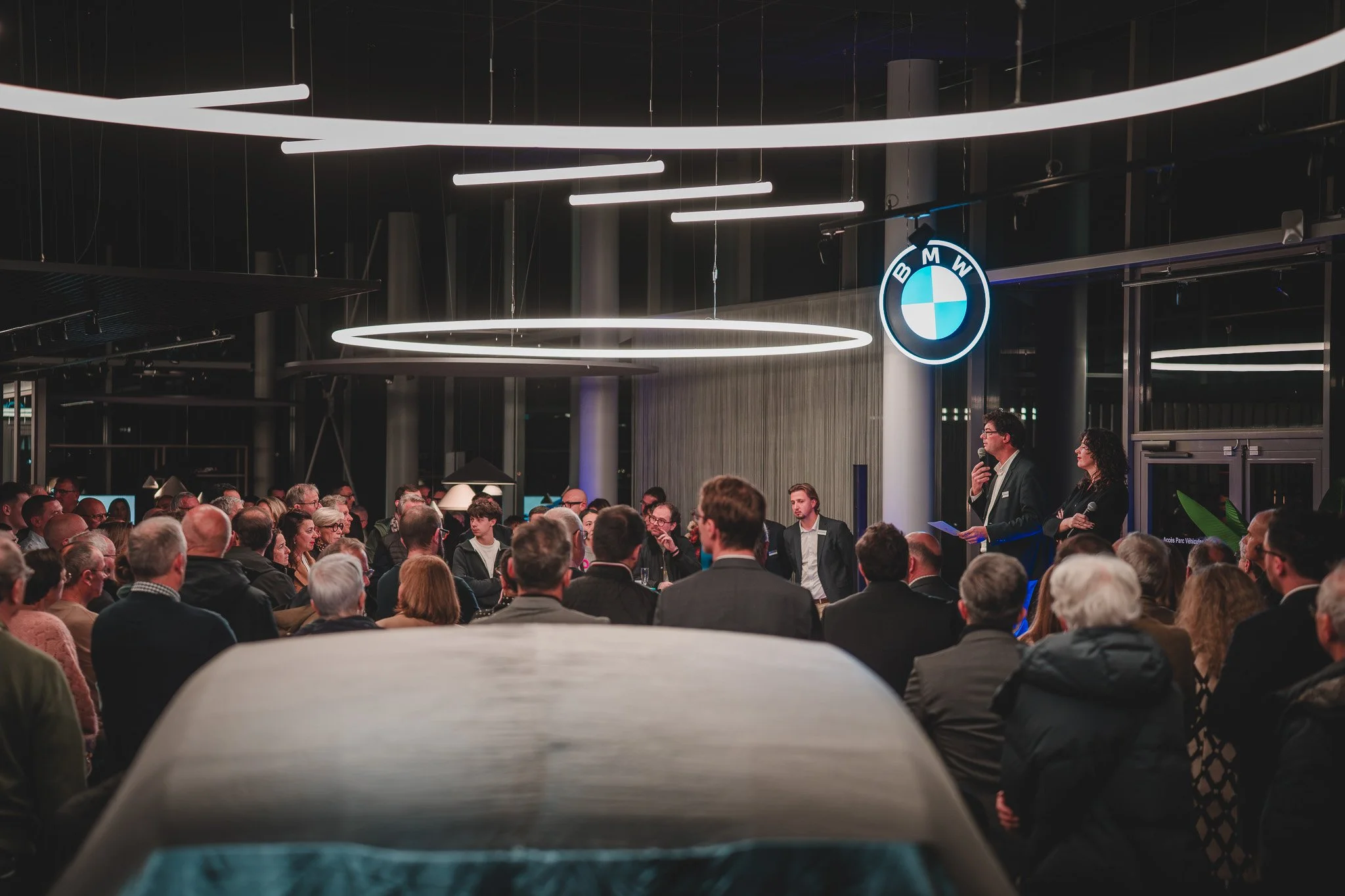 Une réunion ou un événement d'entreprise BMW avec un public nombreux, des personnes qui écoutent une présentation, un logo BMW lumineux en haut à droite.