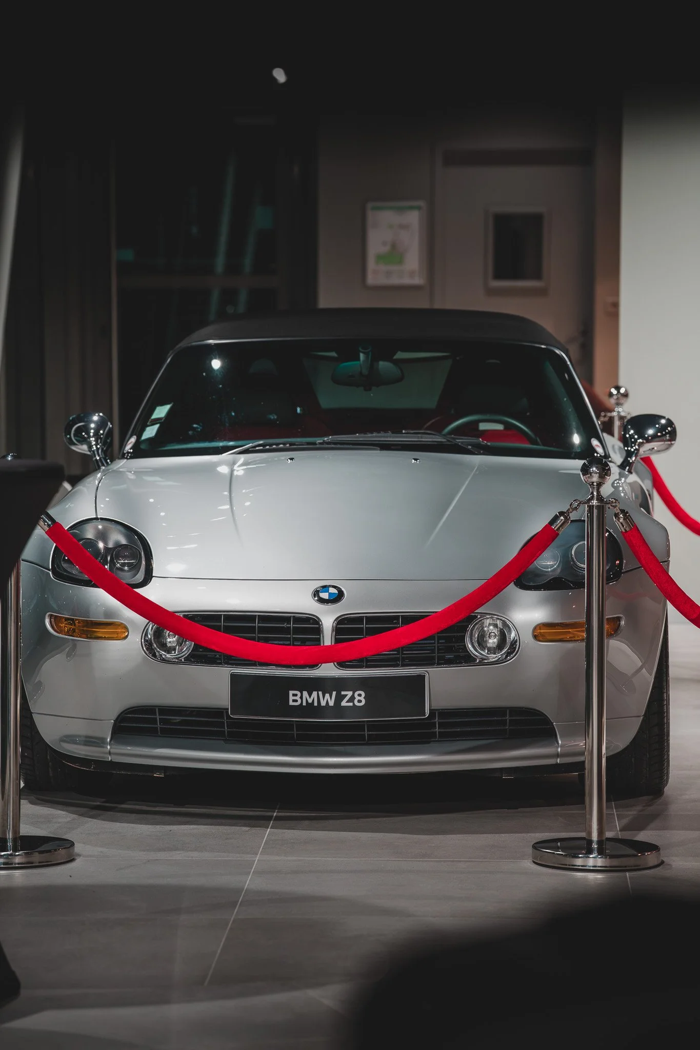 Une voiture BMW Z8 en exposition, entourée de cordes rouges dans un musée ou une galerie, vue de face dans un environnement intérieur