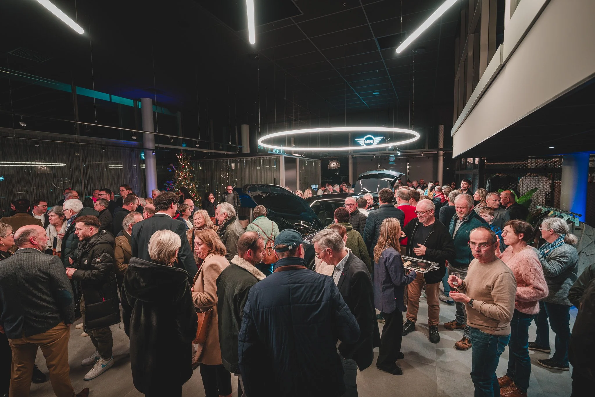 Une foule de personnes à un événement de lancement de voitures dans un showroom. Plusieurs voitures avec capots ouverts sont en exposition. La pièce est décorée pour Noël avec un sapin de Noël et des décorations.