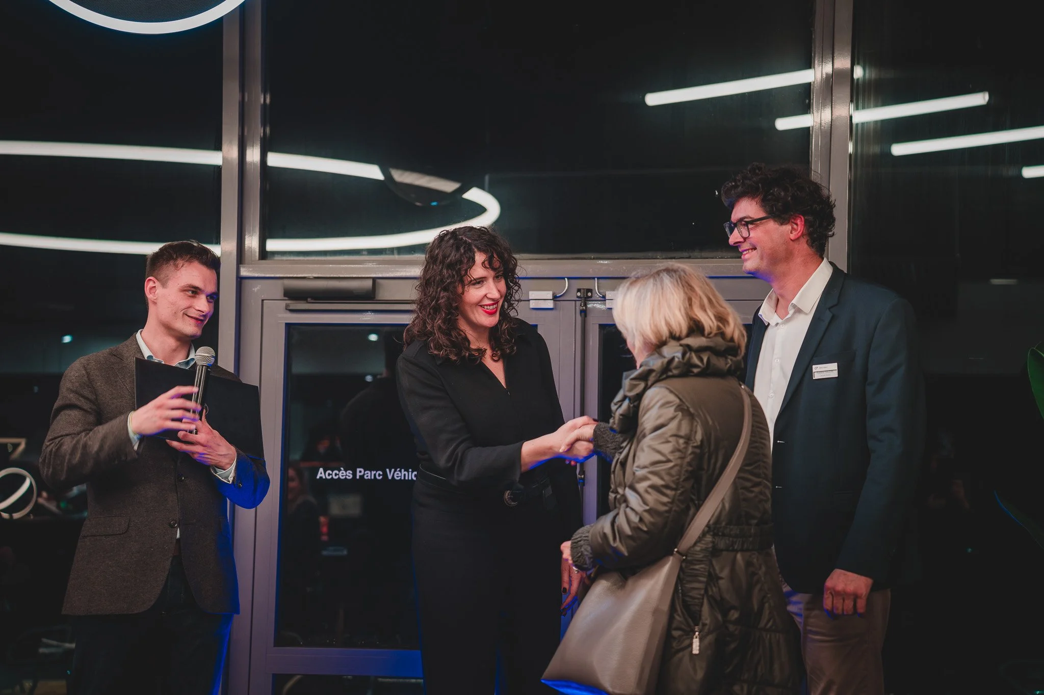 Une femme souriante serre la main d'une femme âgée lors d'une cérémonie d'accueil, entourée de deux hommes. L'un d'eux tient un micro et un dossier, tous dans un environnement intérieur avec un fond sombre et des lumières modernes.