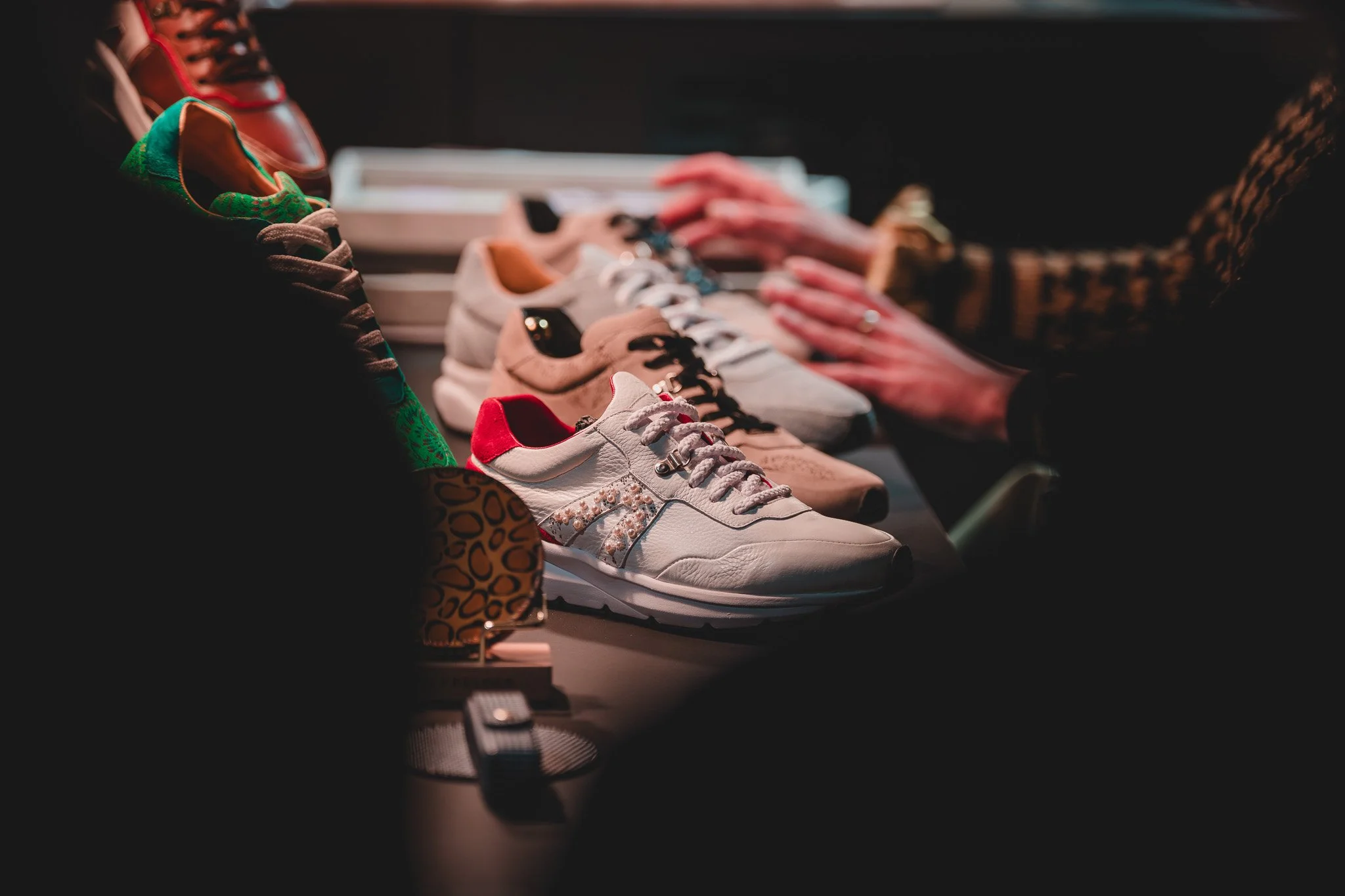 Une sélection de chaussures de différentes couleurs et styles exposées sur une table, avec deux mains d'une personne qui semble faire du shopping en arrière-plan.