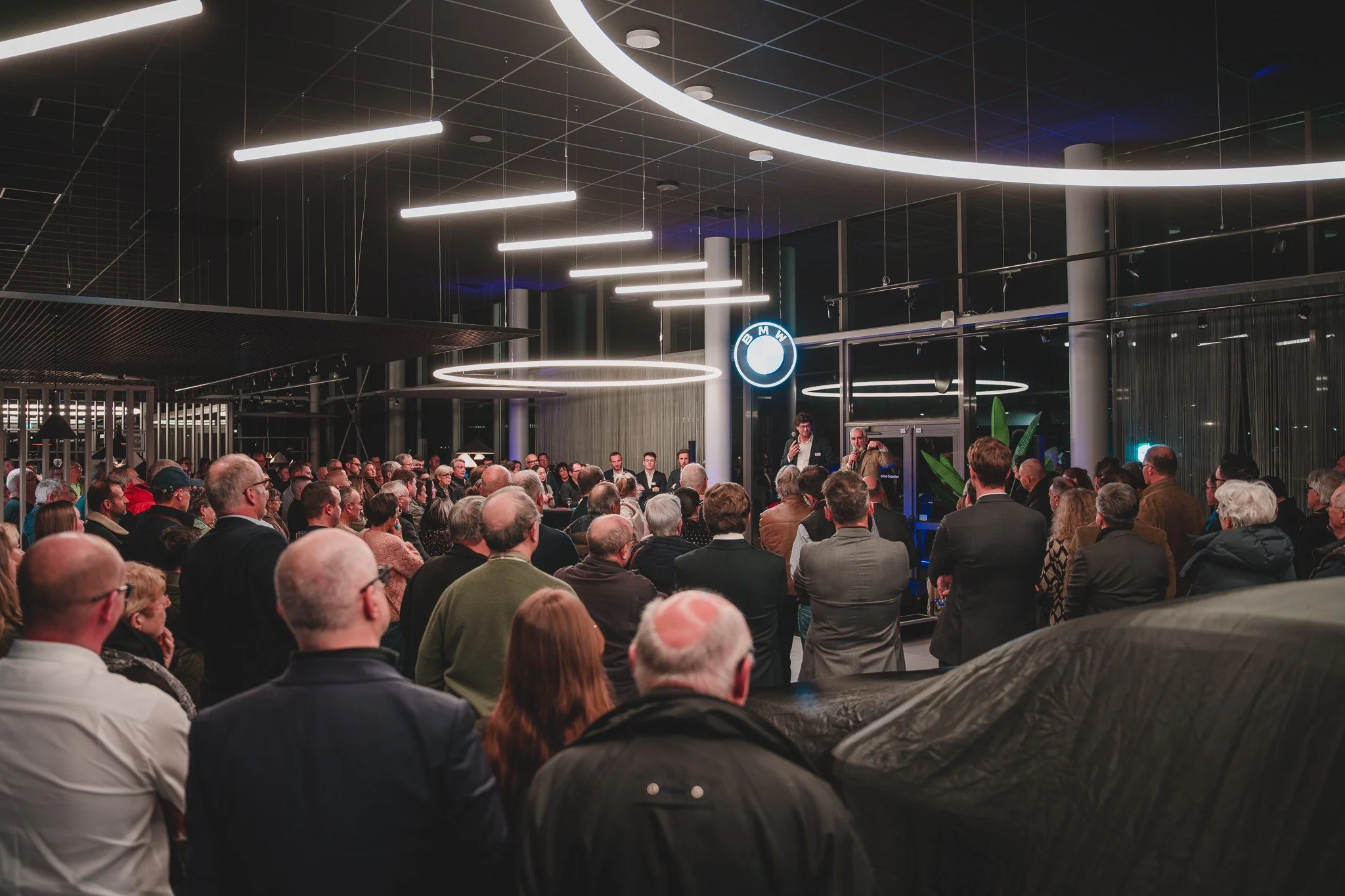 Une assemblée de personnes lors d'une conférence ou d'un événement dans un bâtiment moderne avec un éclairage LED et un logo BMW visible. Les participants sont assis ou debout, écoutant deux orateurs en avant.