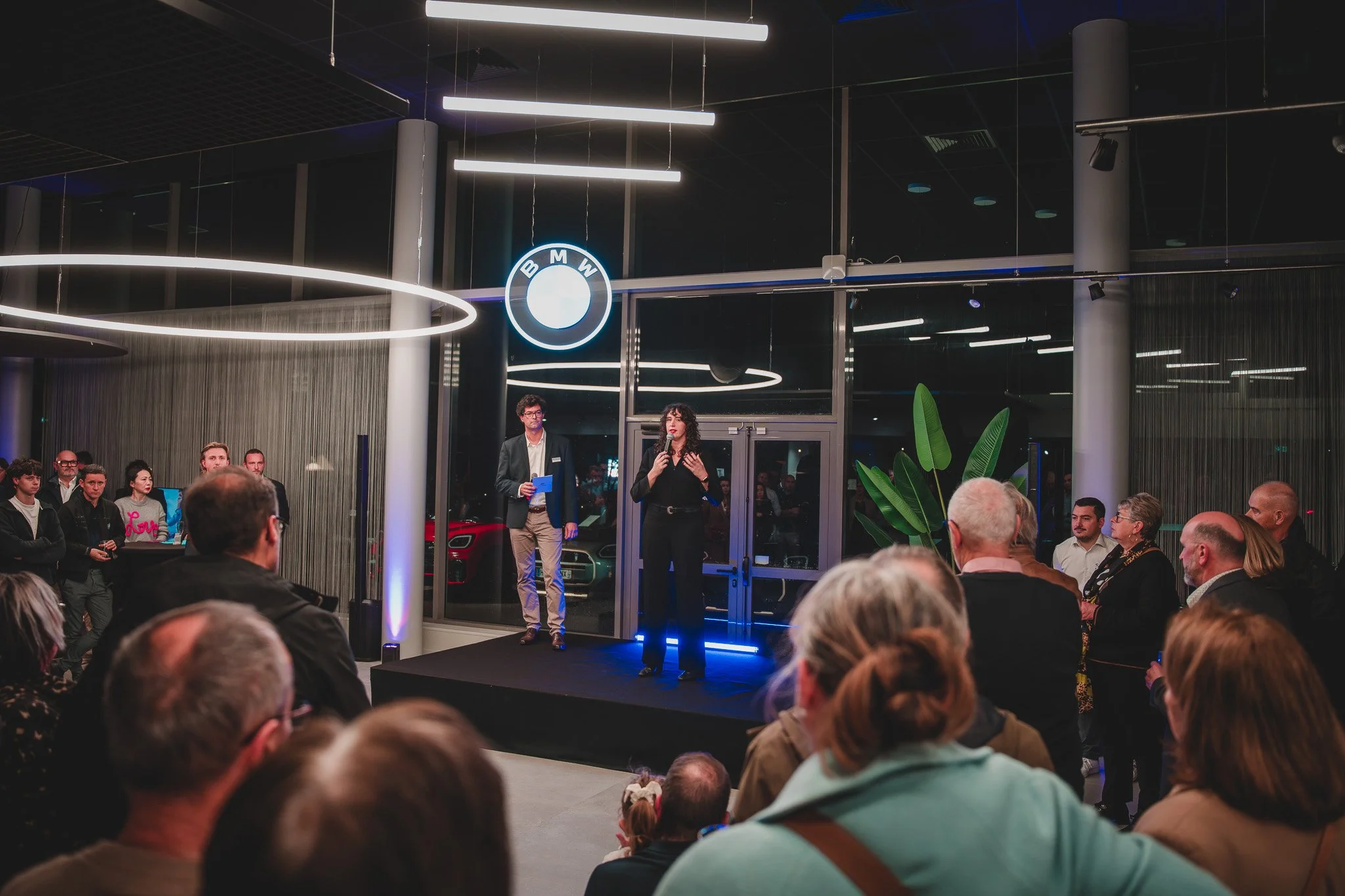 Une cérémonie ou un événement de dévoilement dans un showroom BMW, avec un public rassemblé, un homme et une femme parlant sur une petite scène devant une grande vitrine, un logo BMW illuminé au mur, et une voiture visible à l'extérieur.