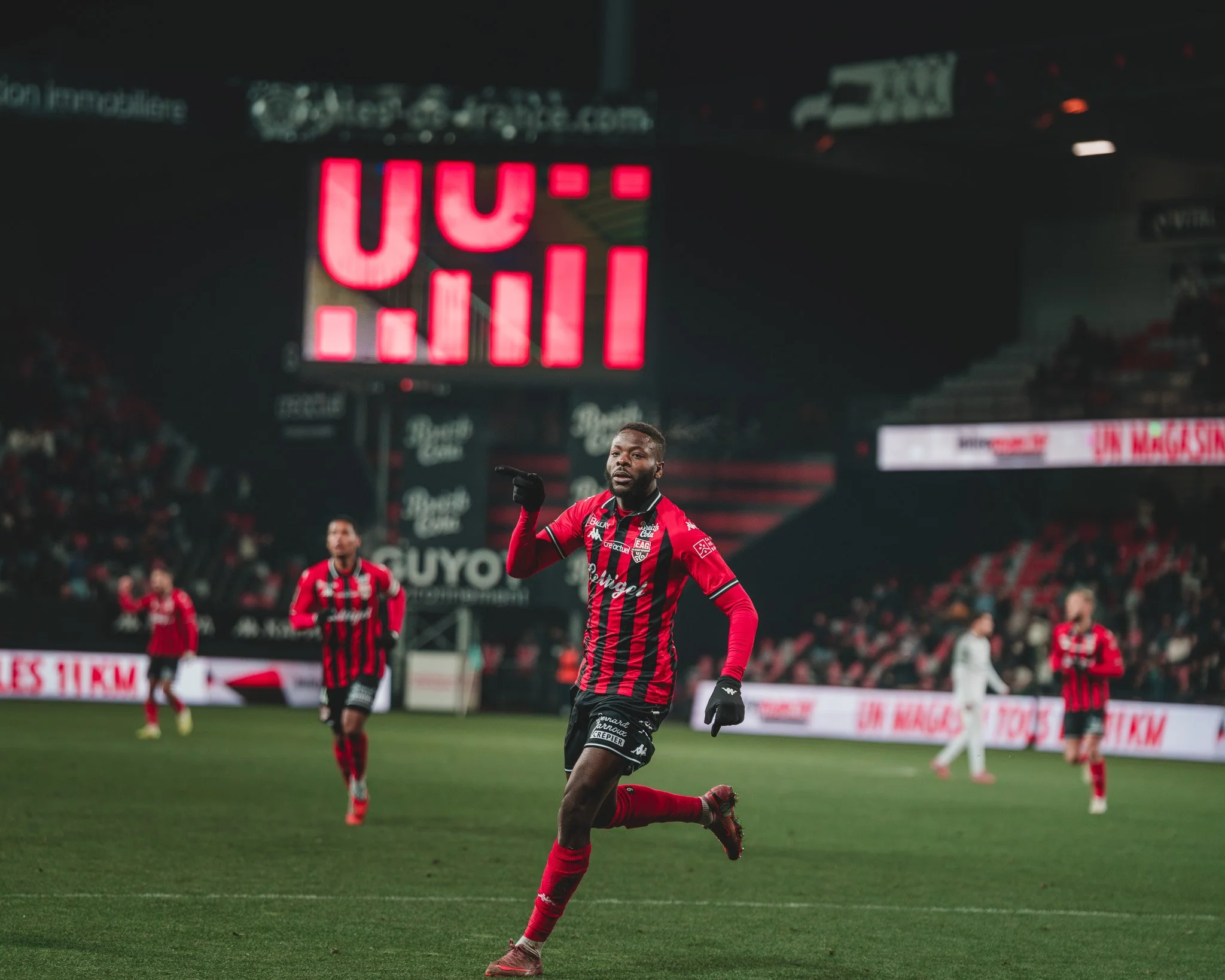 Un joueur de football en train de courir sur le terrain lors d'un match, portant un maillot rouge et noir, avec d'autres joueurs en arrière-plan dans un stade.