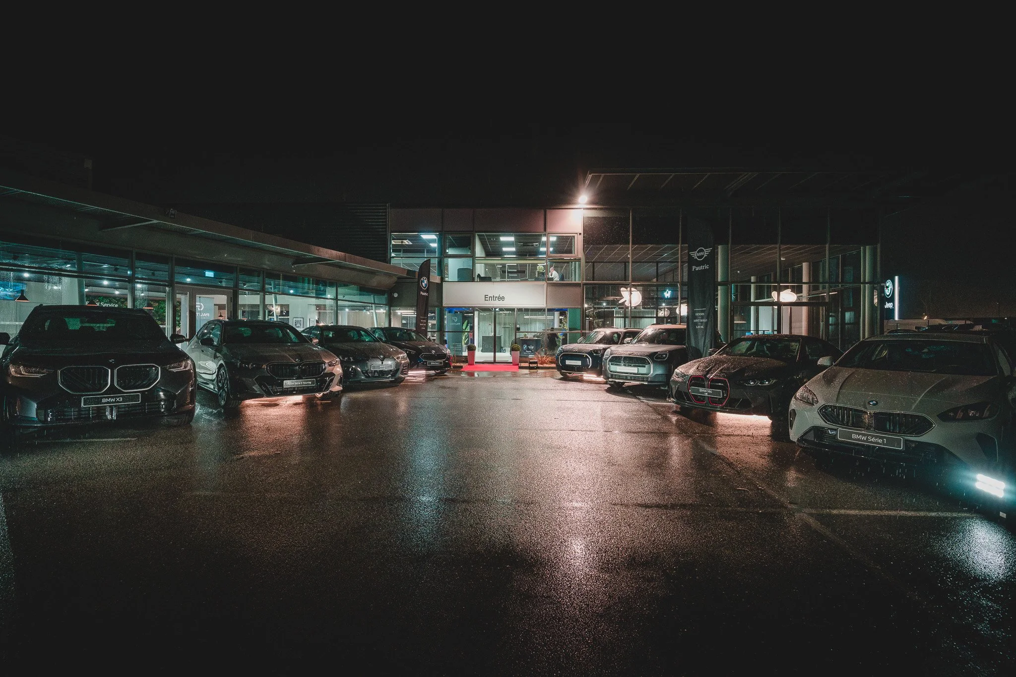 Vue de l'entrée d'un concessionnaire automobile avec plusieurs voitures modernes alignées, éclairée la nuit avec un sol mouillé reflétant les lumières.