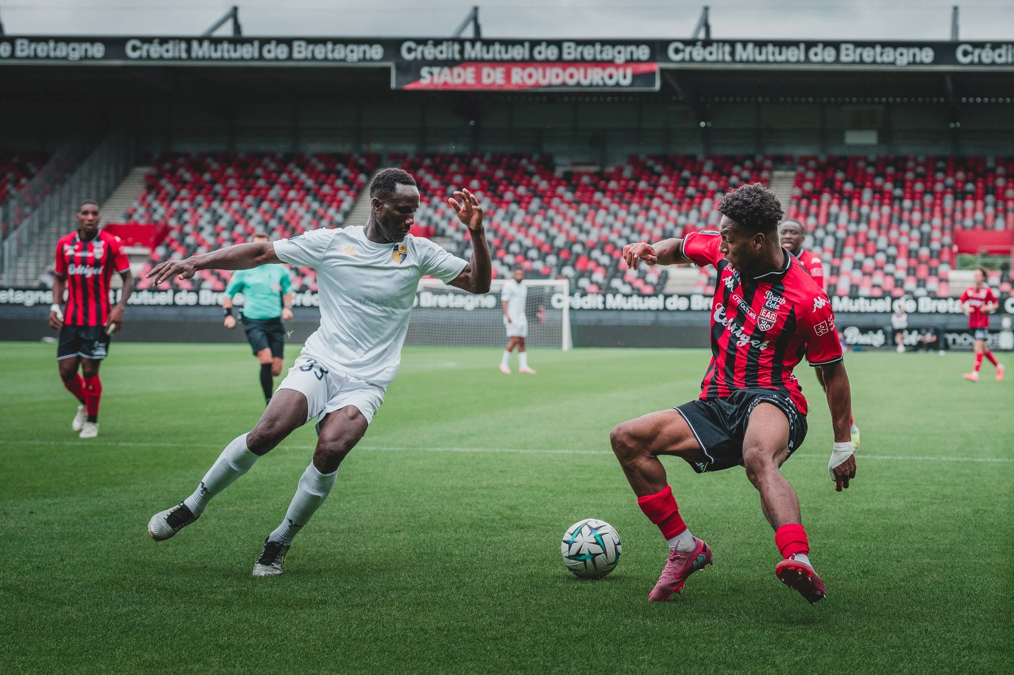 Deux joueurs de football en action sur le terrain, un en maillot blanc et l'autre en maillot rouge et noir, lors d'un match au stade de Roudourou.