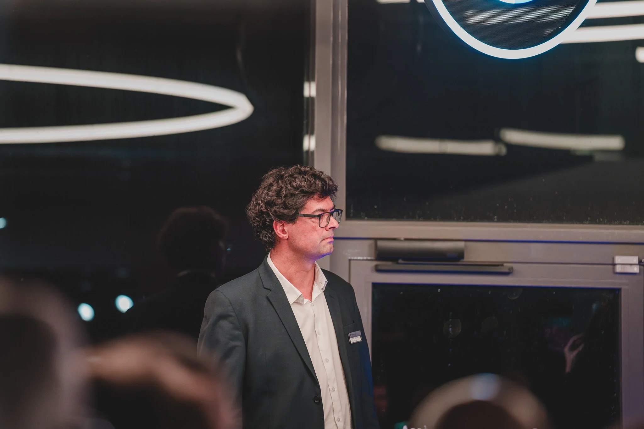 Un homme avec des cheveux bouclés et portant des lunettes, en costume sombre, regarde fixement à côté, dans un lieu sombre avec des lumières en arrière-plan.