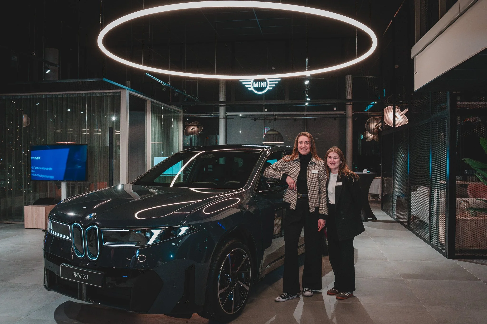 Deux femmes posant devant une voiture BMW iX3 dans un showroom, avec un éclairage moderne et un logo MINI suspendu au plafond.
