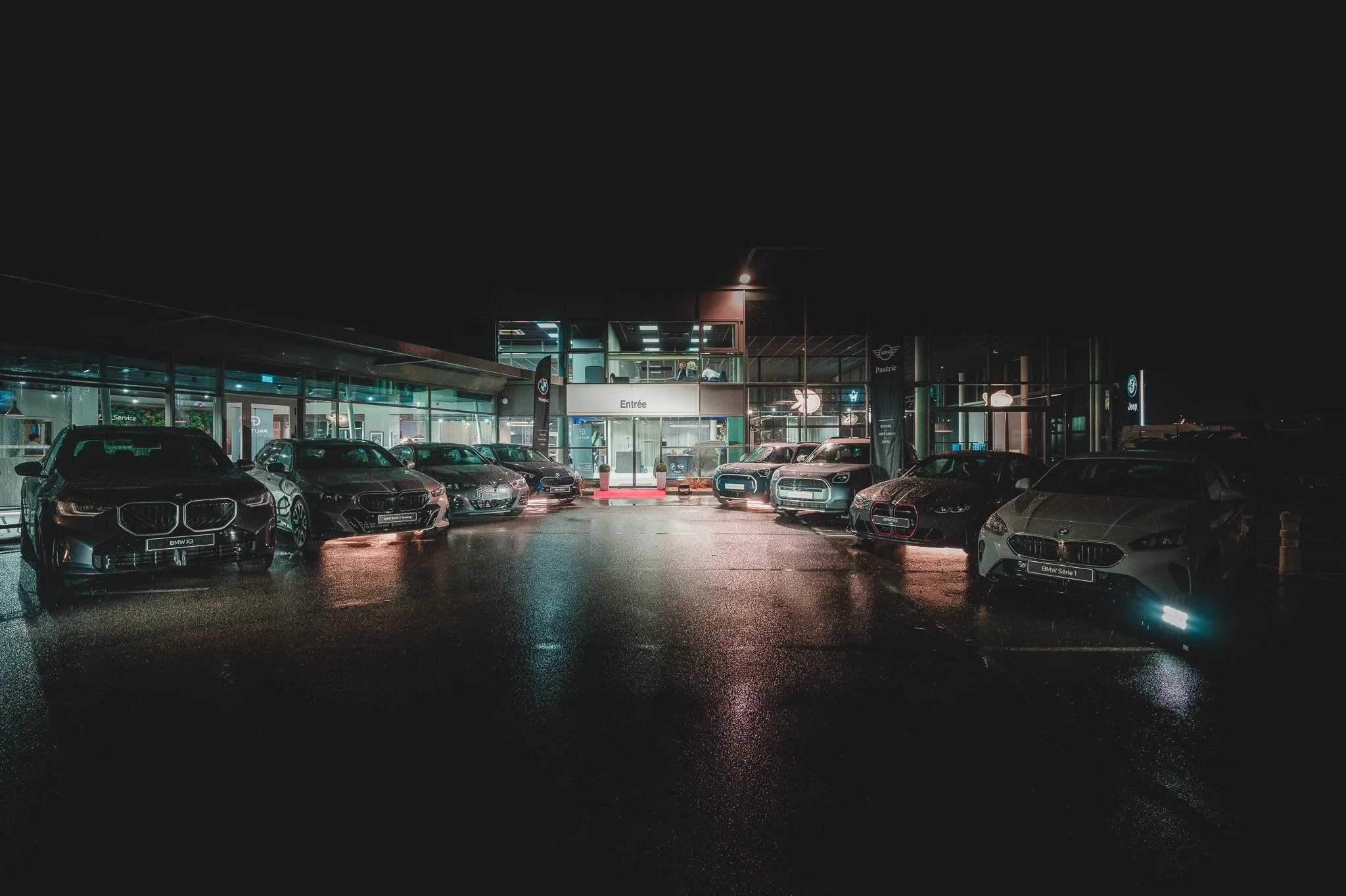 Parking éclairé devant un concessionnaire automobile la nuit, avec plusieurs voitures modernes de différentes marques alignées.