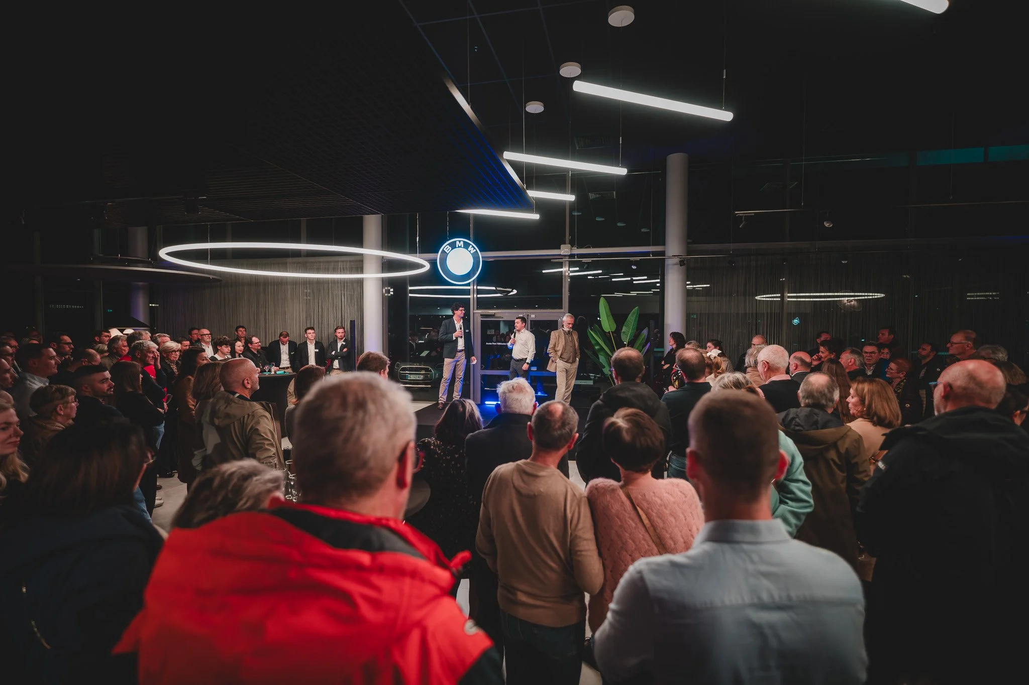 Une réunion d'affaires ou un événement social dans un espace moderne avec un public nombreux. Un homme parle sur scène avec un micro, entouré de plusieurs autres personnes. Le décor inclut un logo BMW brillant au-dessus de la scène.