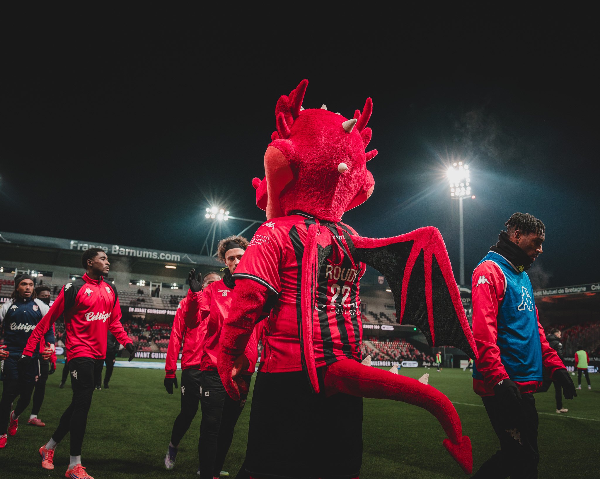 Une mascotte de démon rouge avec des cornes, des ailes, et une queue, portant un maillot de football, dans un stade de football la nuit, entourée de joueurs. Le décor est illuminé par des projecteurs.