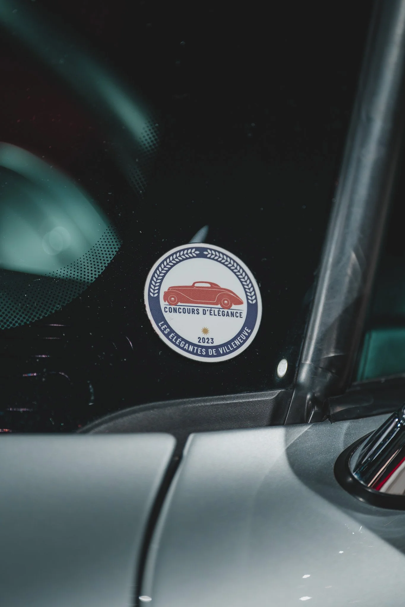 Autocollant ronds sur une voiture, avec une illustration d'une voiture ancienne en rouge et des textes en français, mentionnant un concours d'élégance pour l'édition 2023.