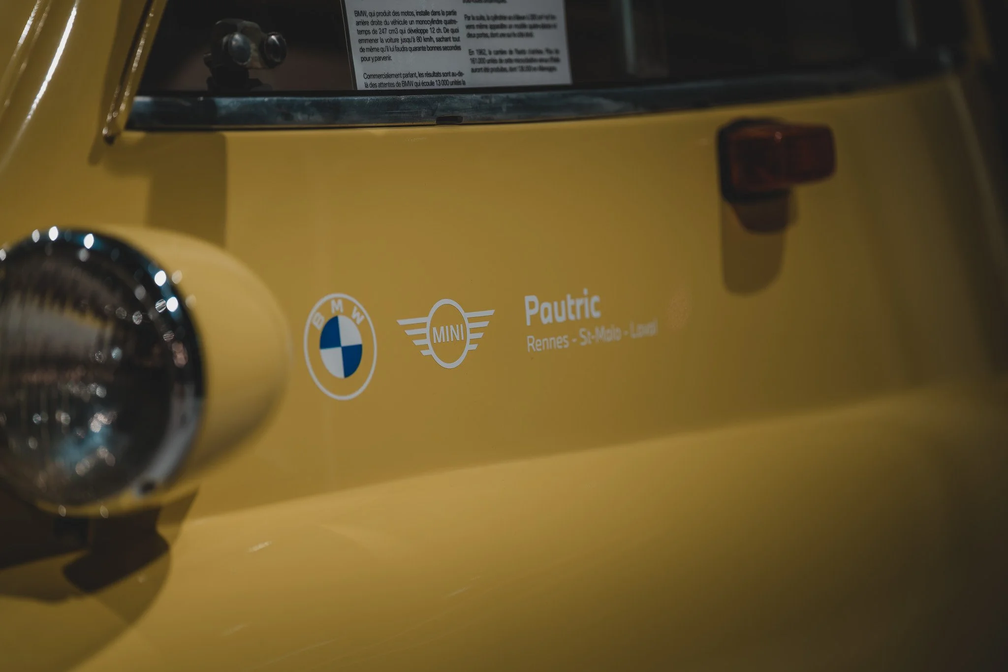 Détail de la carrosserie d'une miniature de voiture jaune avec logos BMW et MINI, et le nom 'Pautric' accompagné de localisations 'Rennes, Saint-Malo, Léon'.