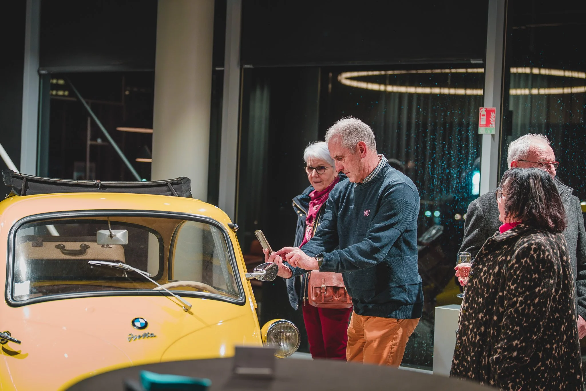 Groupe de personnes regardant une petite voiture jaune BMW lors d'une exposition ou d'un événement à l'intérieur.