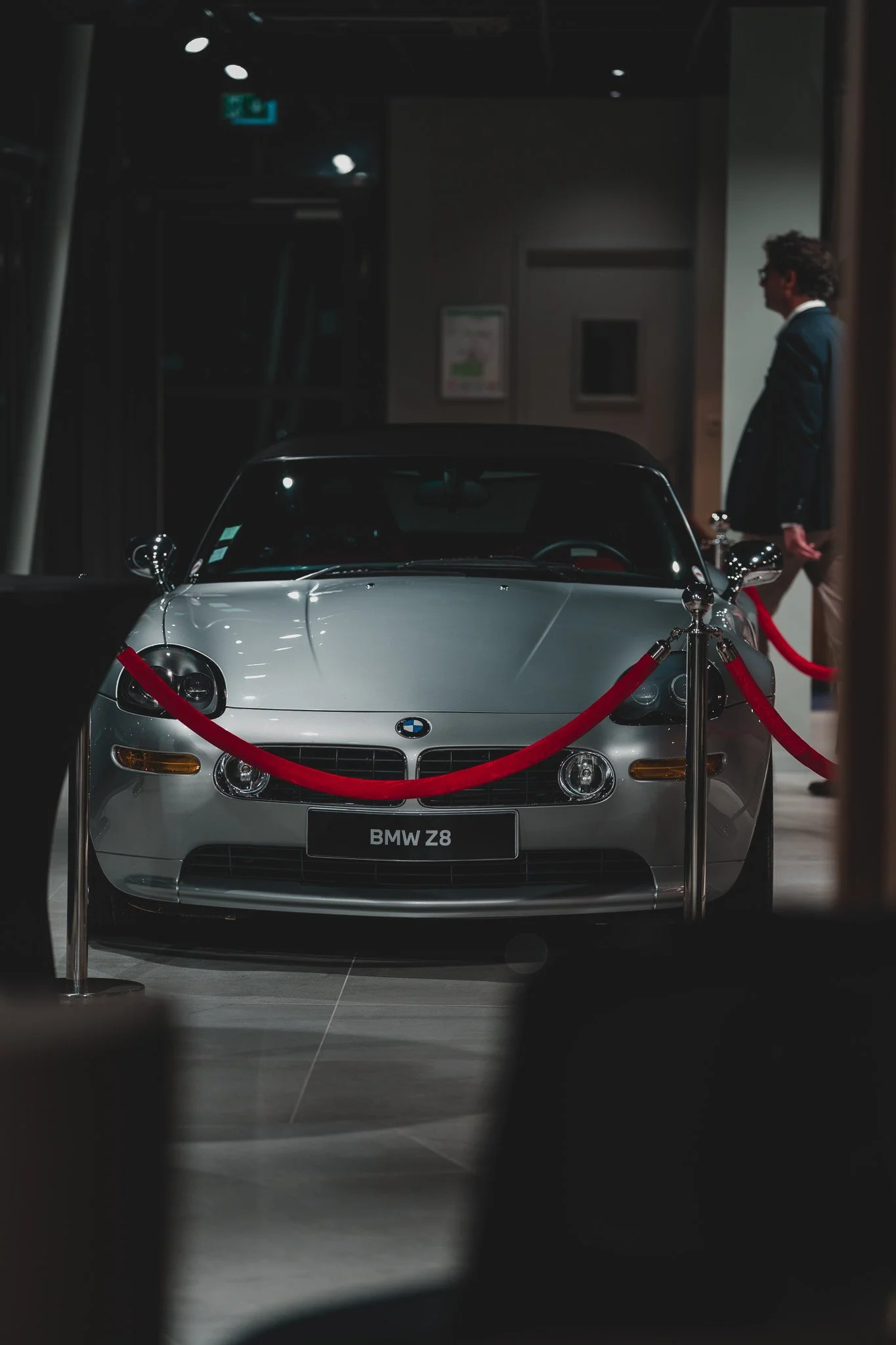Une voiture BMW Z8 exposée derrière une barrière de velours rouge dans une salle d'exposition, avec une personne en costume à l'arrière-plan.