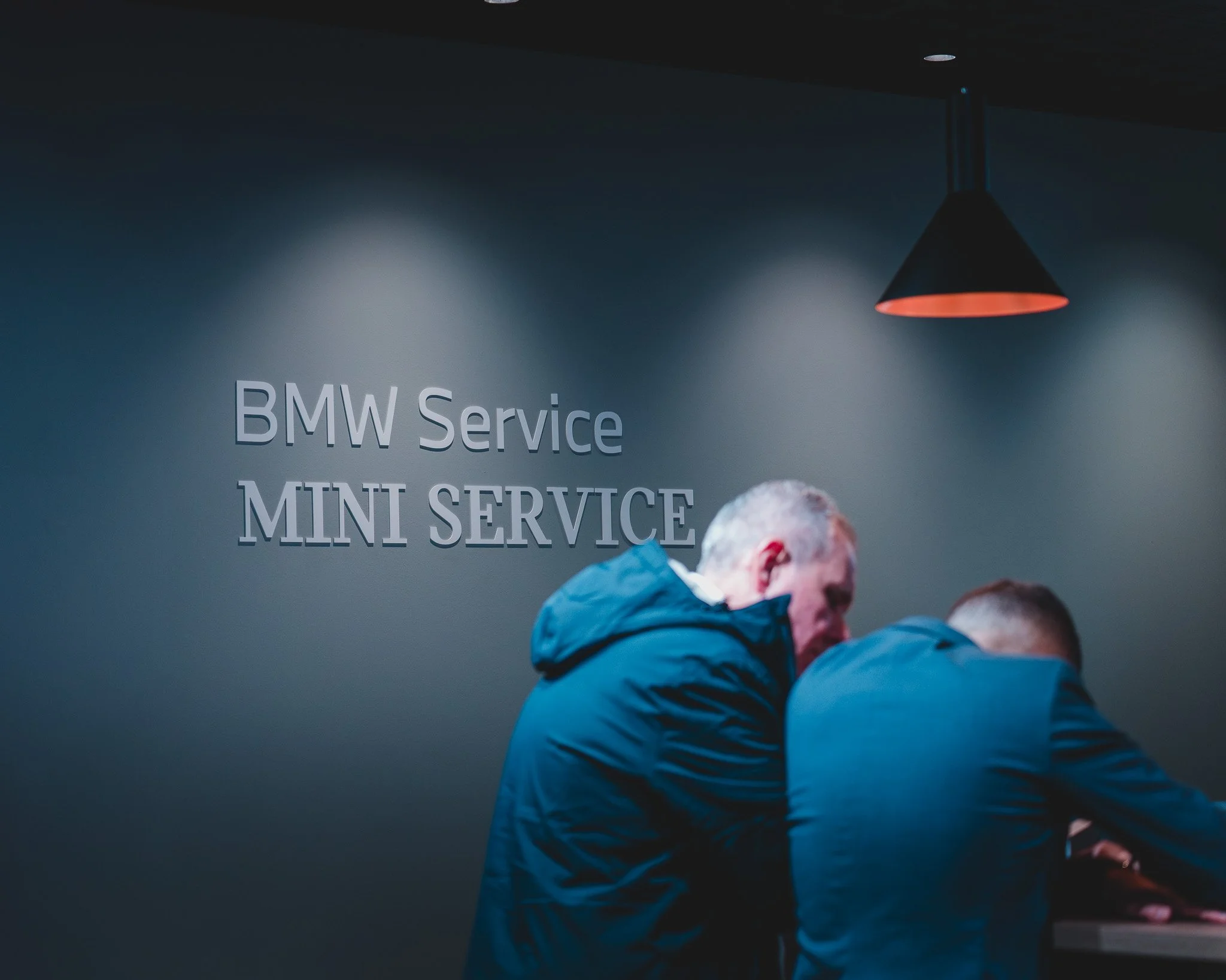 Deux hommes assis au comptoir du service BMW/MINI, un éclairage tamisé et un panneau sur le mur indiquant 'BMW Service MINI SERVICE'.