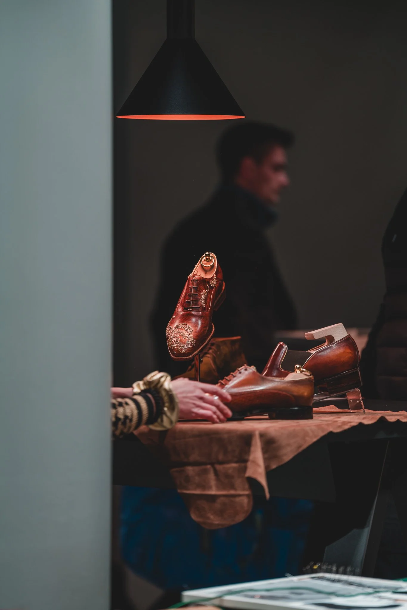 Gants, chaussures en cuir et une chaussure suspendue sur un présentoir en vente dans une boutique, en arrière-plan une personne floue et un luminaire
